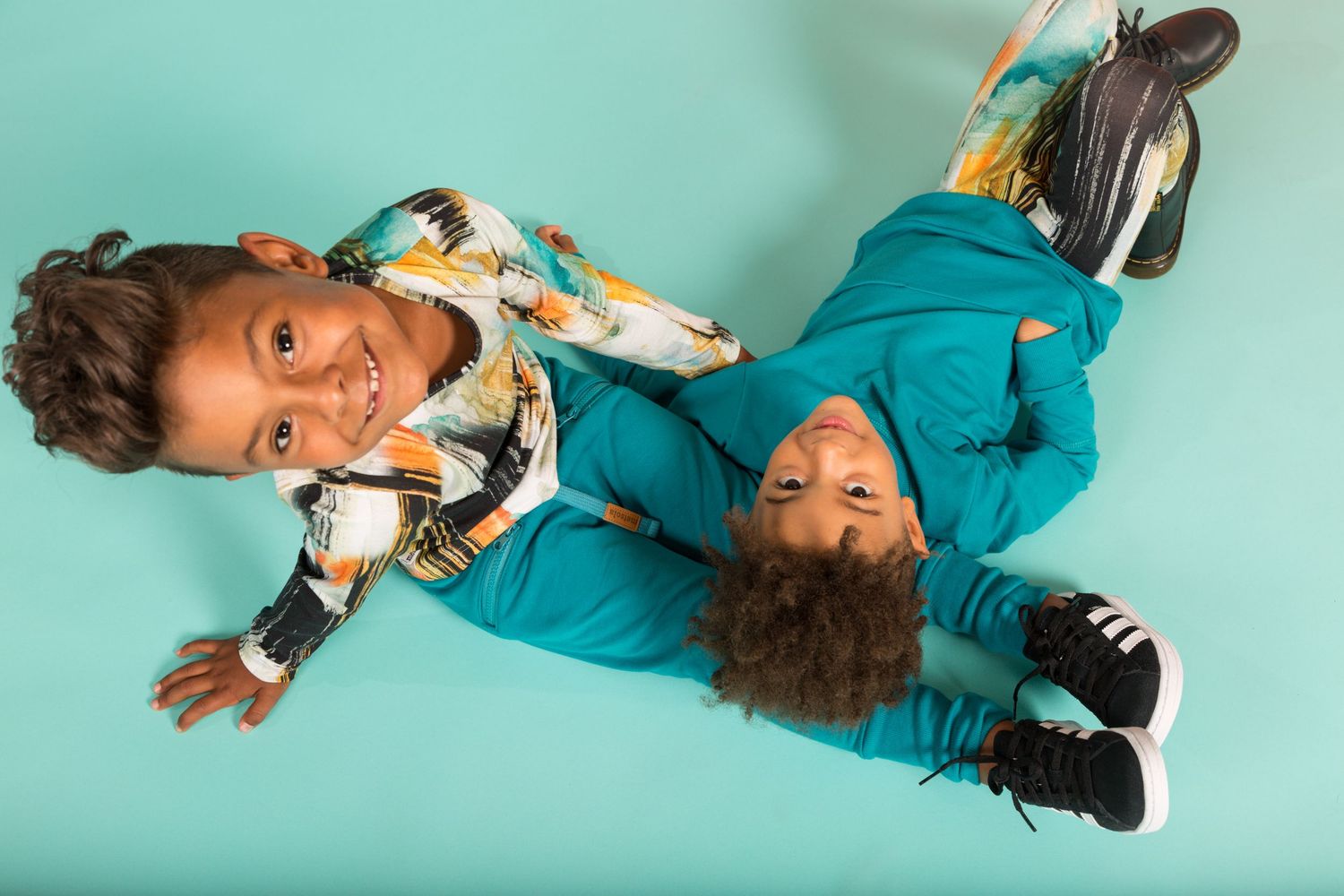 Two children in colorful clothing lay on a turquoise background while posing playfully for the camera.