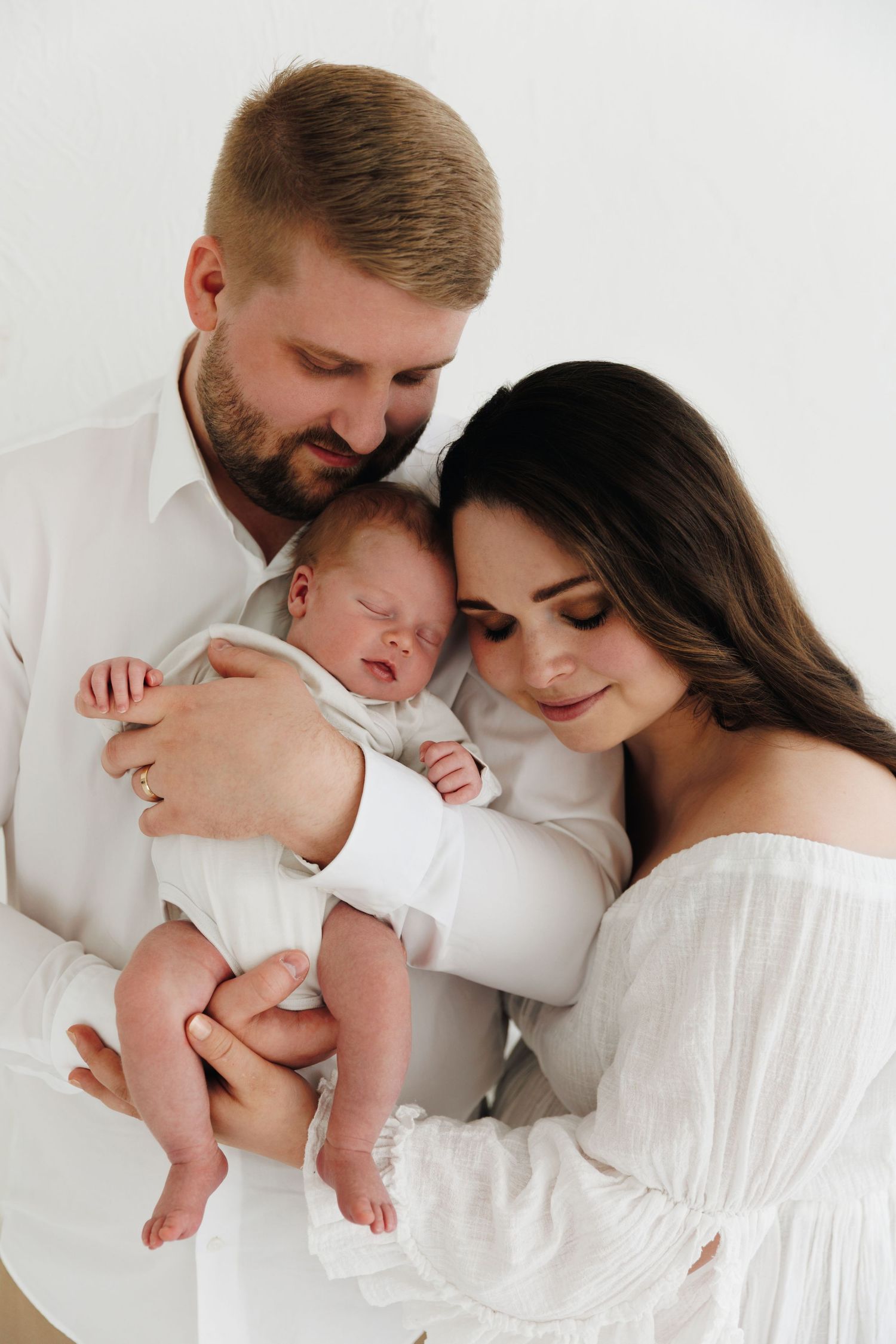 A couple in white outfits tenderly holds their sleeping newborn baby in a bright, minimalist studio setting.