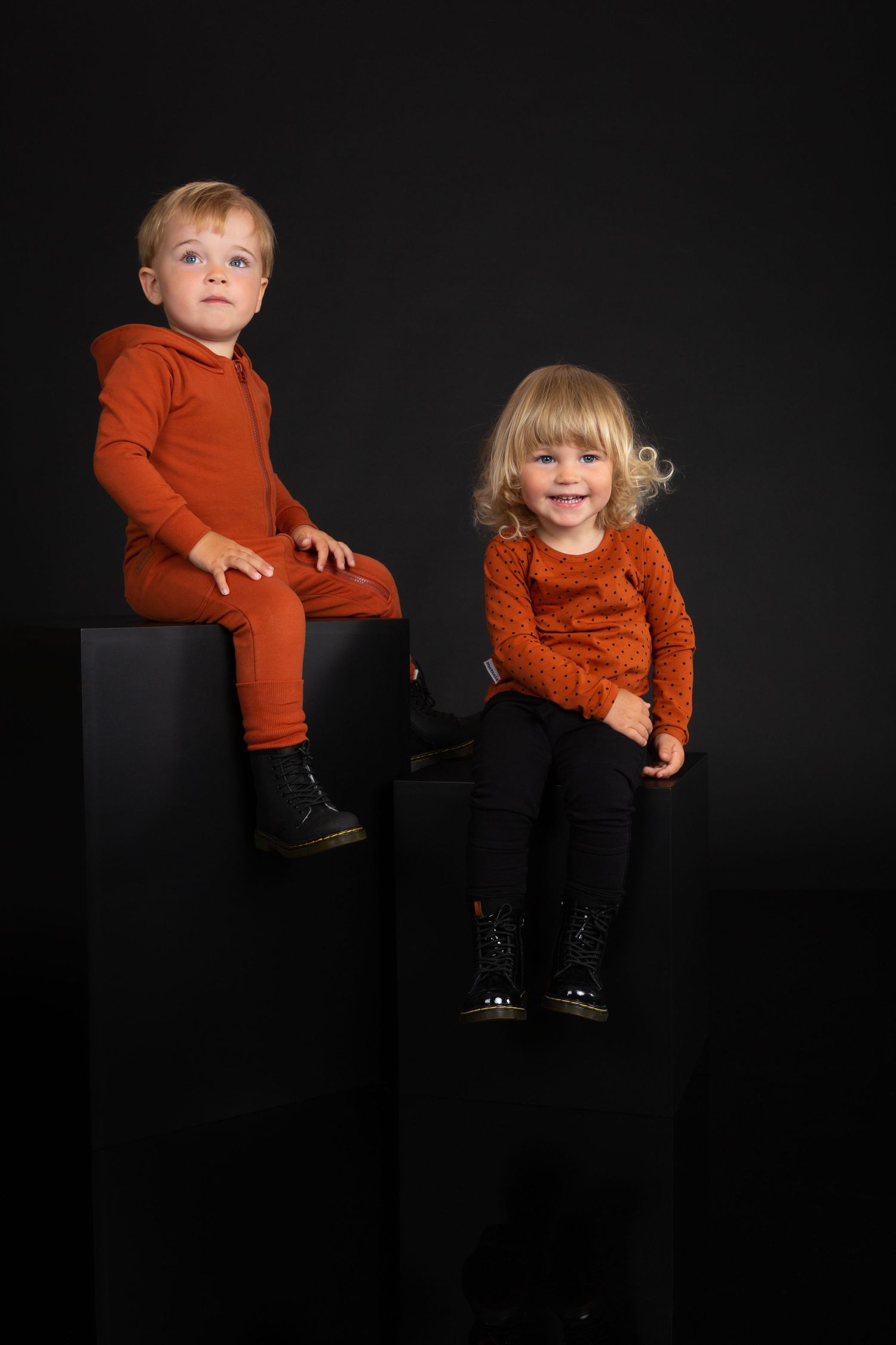 Two young blonde children in matching rust-orange outfits sit on black cubes against a dark studio background.
