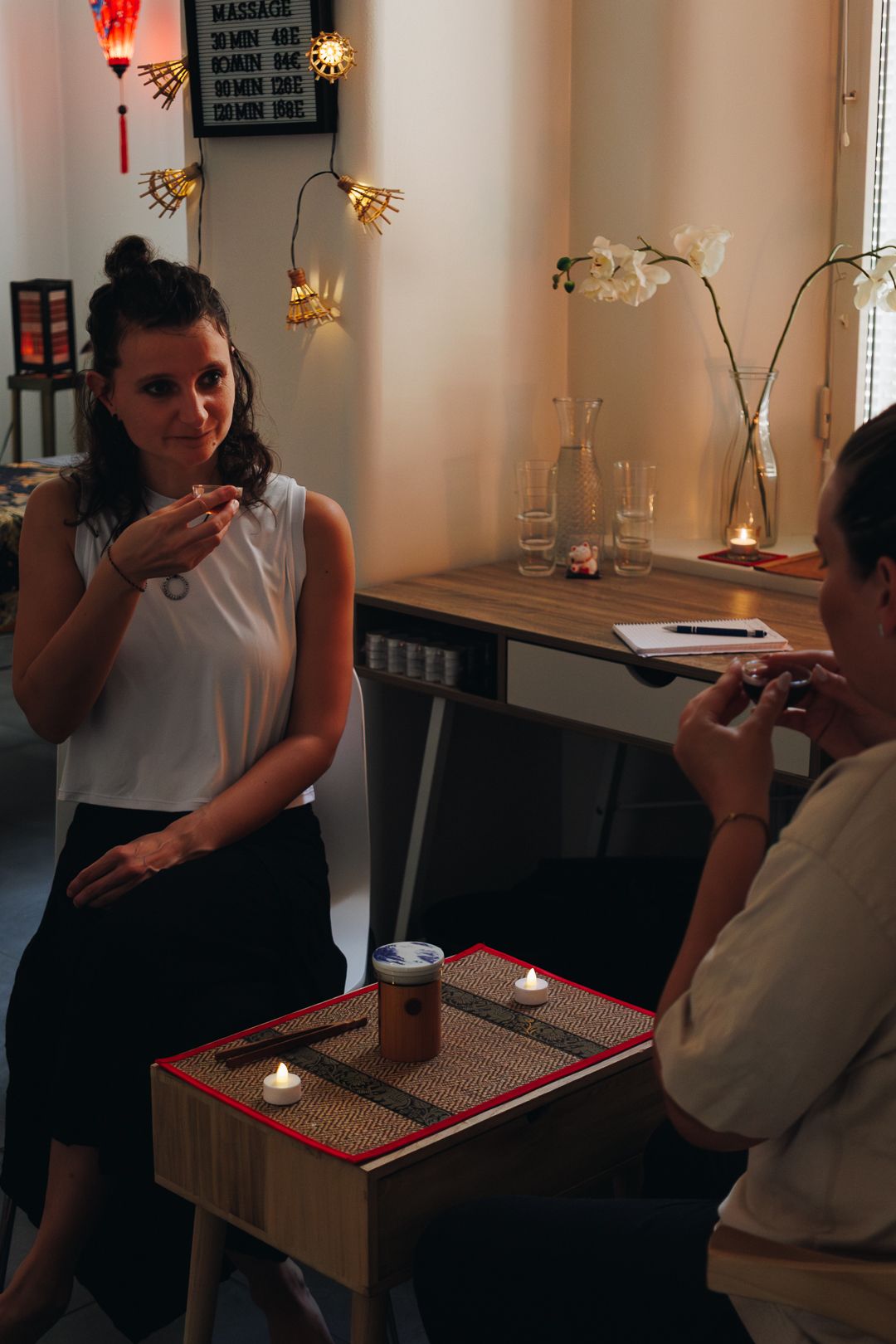Two women enjoying a traditional tea ceremony at a candlelit table in a cozy wellness studio.