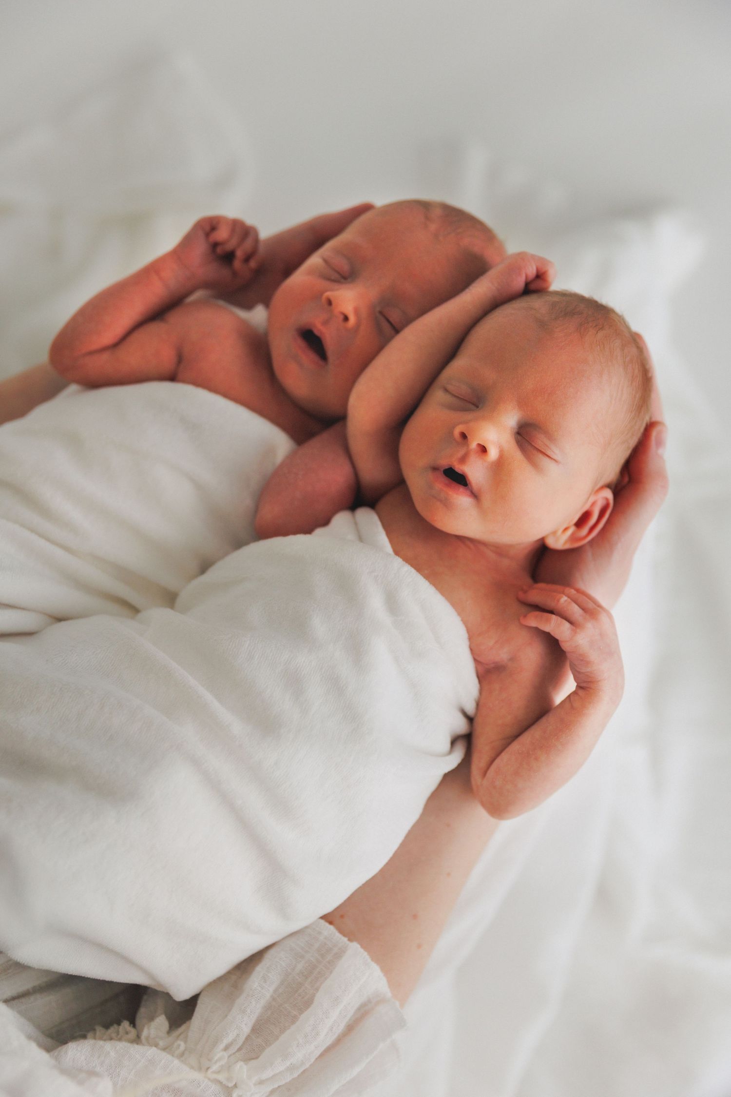 Two sleeping newborn twins lying together on a white blanket, cradled by an adult hand.