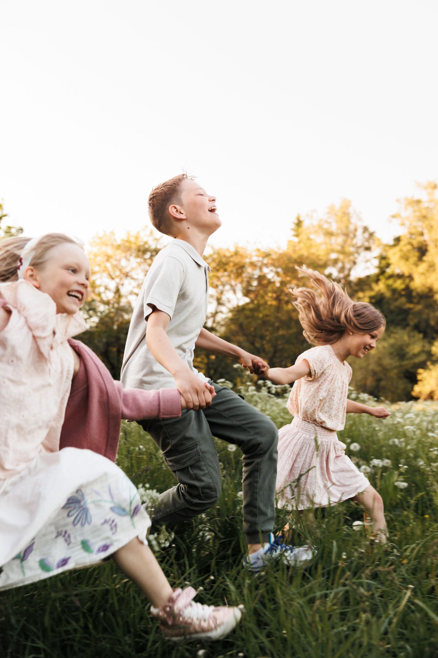 Three happy children running and holding hands through a sunny wildflower meadow, laughing joyfully outdoors.