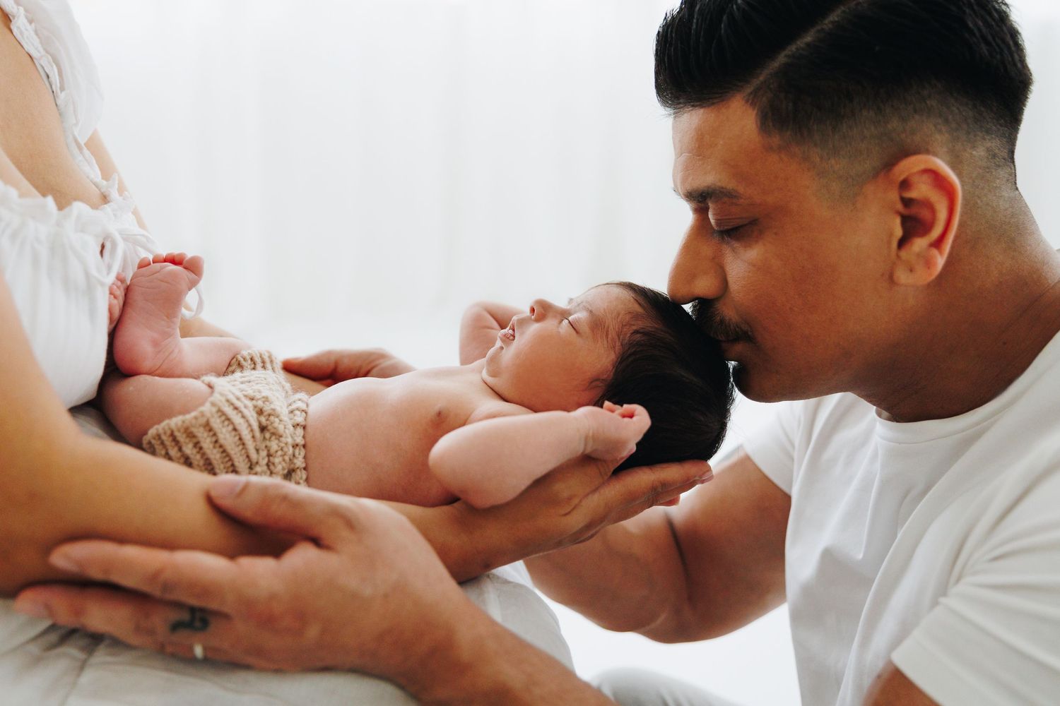 A father tenderly kisses his newborn baby held by the mother in a bright, serene setting.