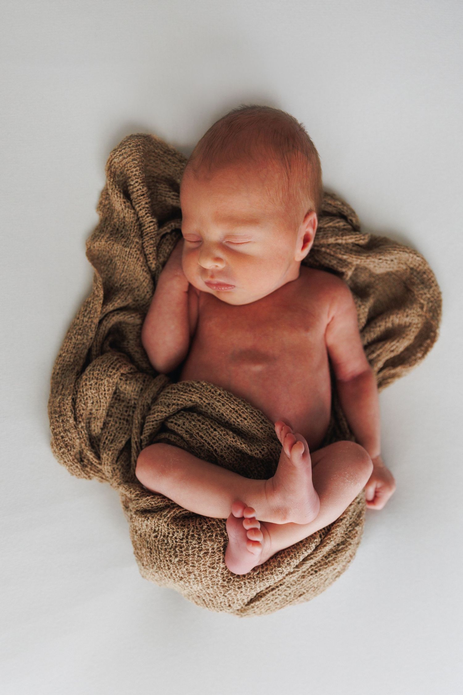 Peaceful newborn baby sleeping curled up on a soft brown knit blanket against a white background.