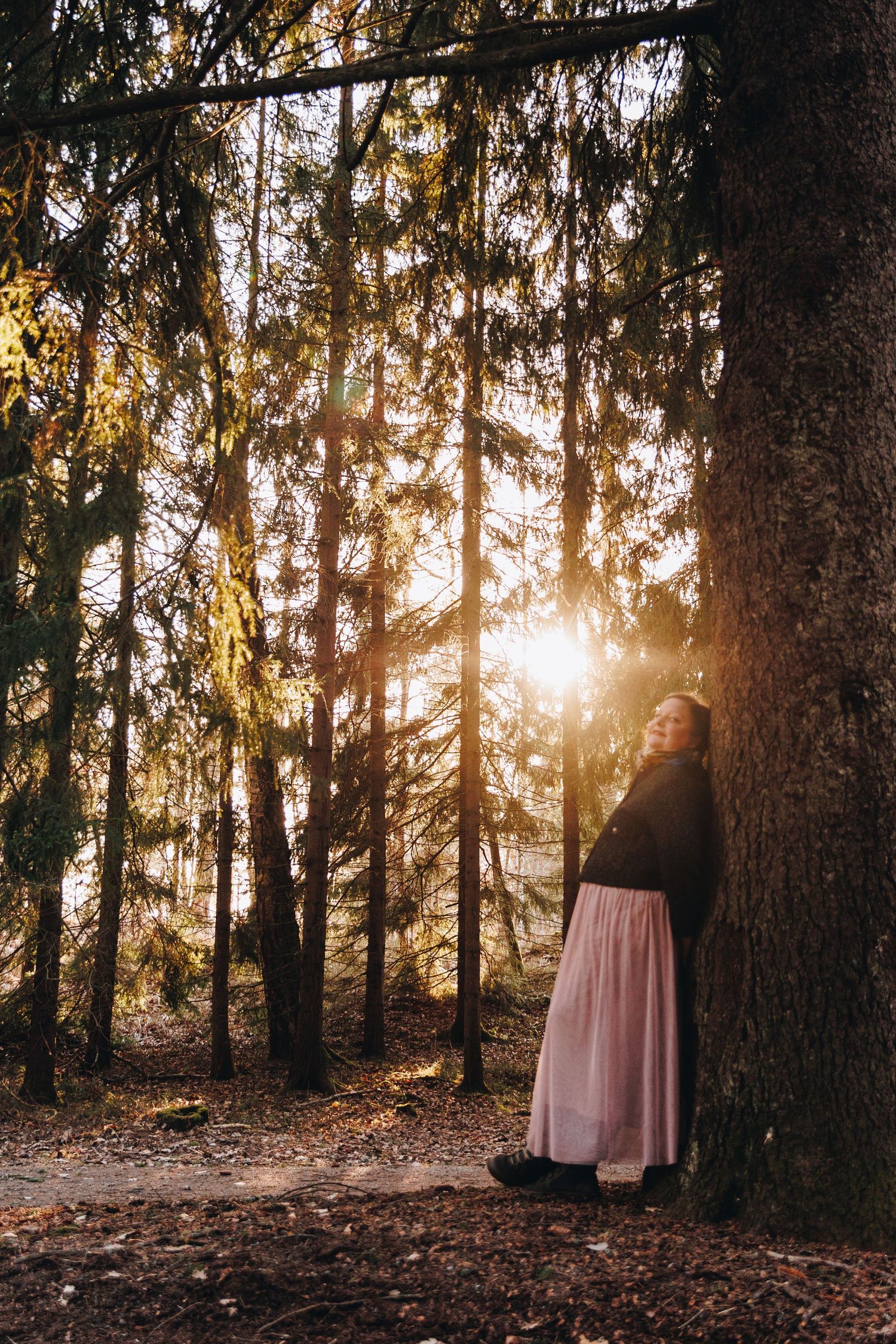Woman in pink skirt leaning against pine tree in sunlit forest at golden hour.