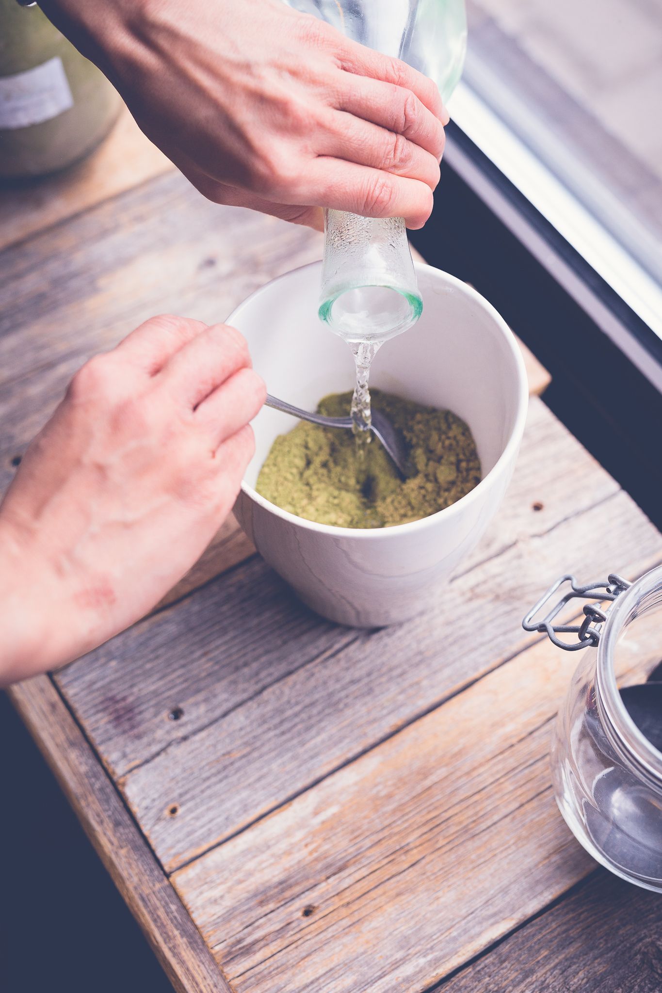 Hands pouring water into a white bowl with green matcha paste on a rustic wooden surface.