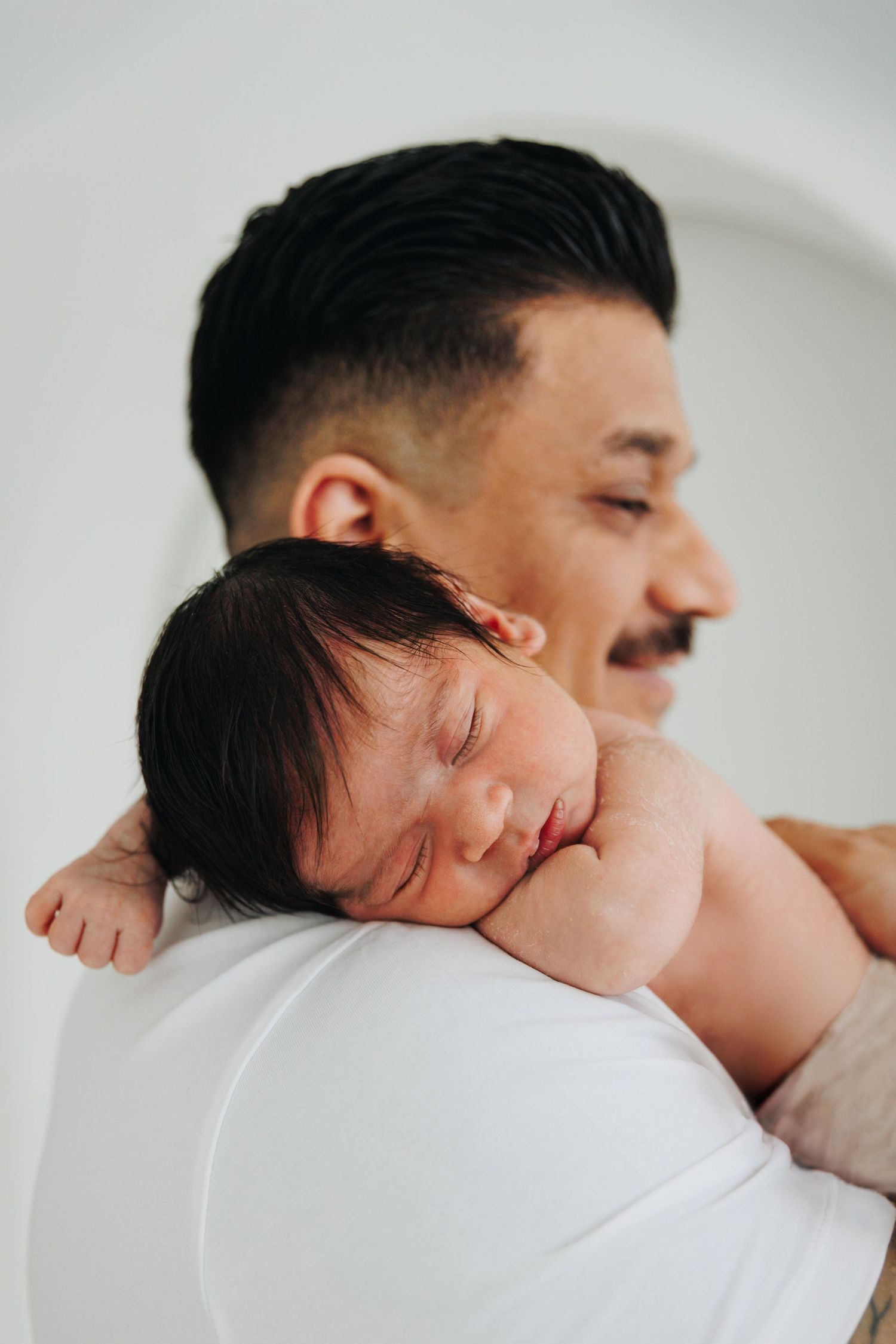 A smiling father with a mustache gently holds his sleeping newborn baby on his shoulder.