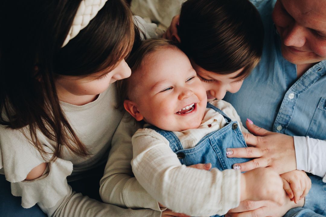 A laughing baby plays and cuddles with parents on a white bed while wearing cozy knitwear.