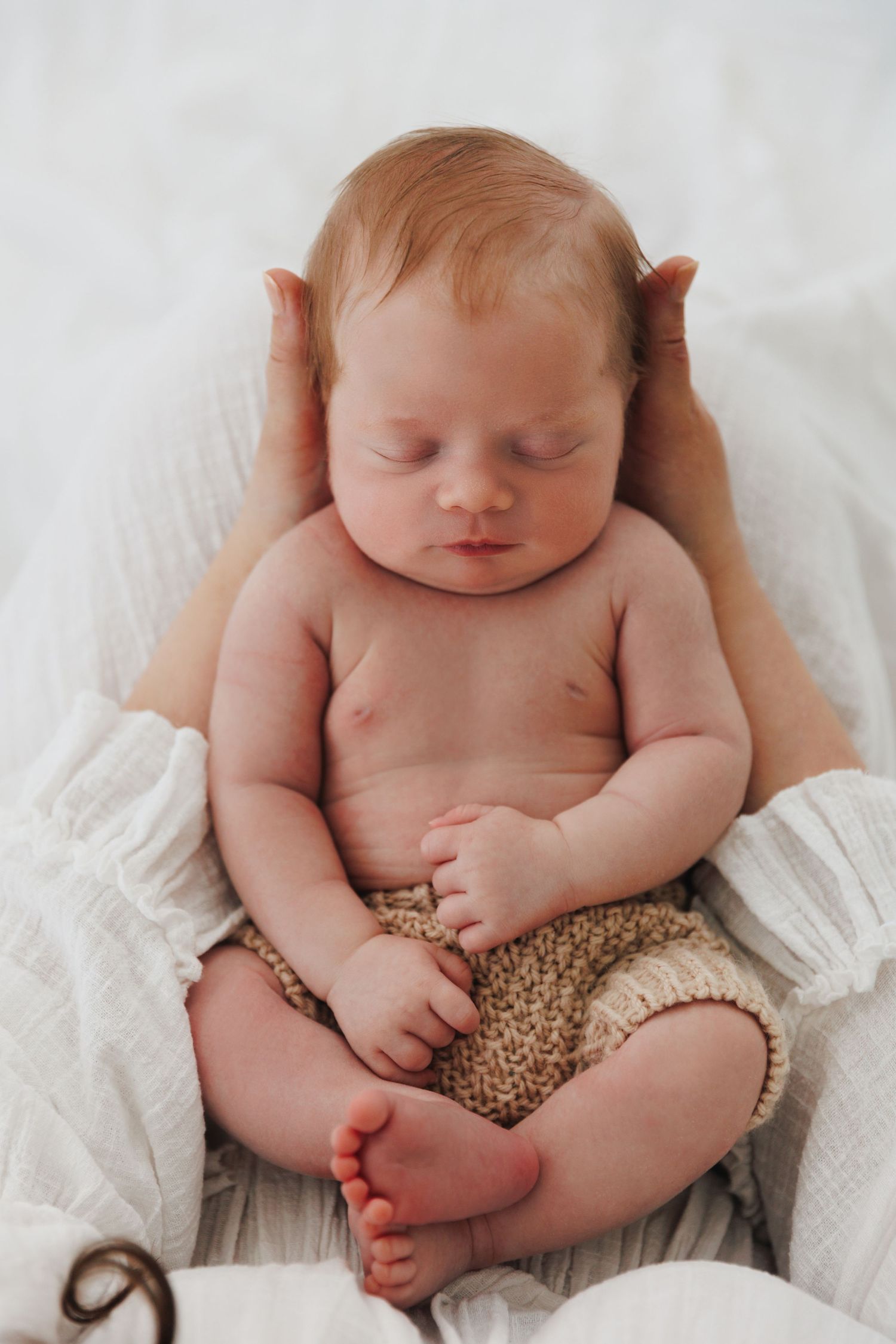 Sleeping newborn baby with red hair wearing knit shorts, cradled in adult hands on white fabric.