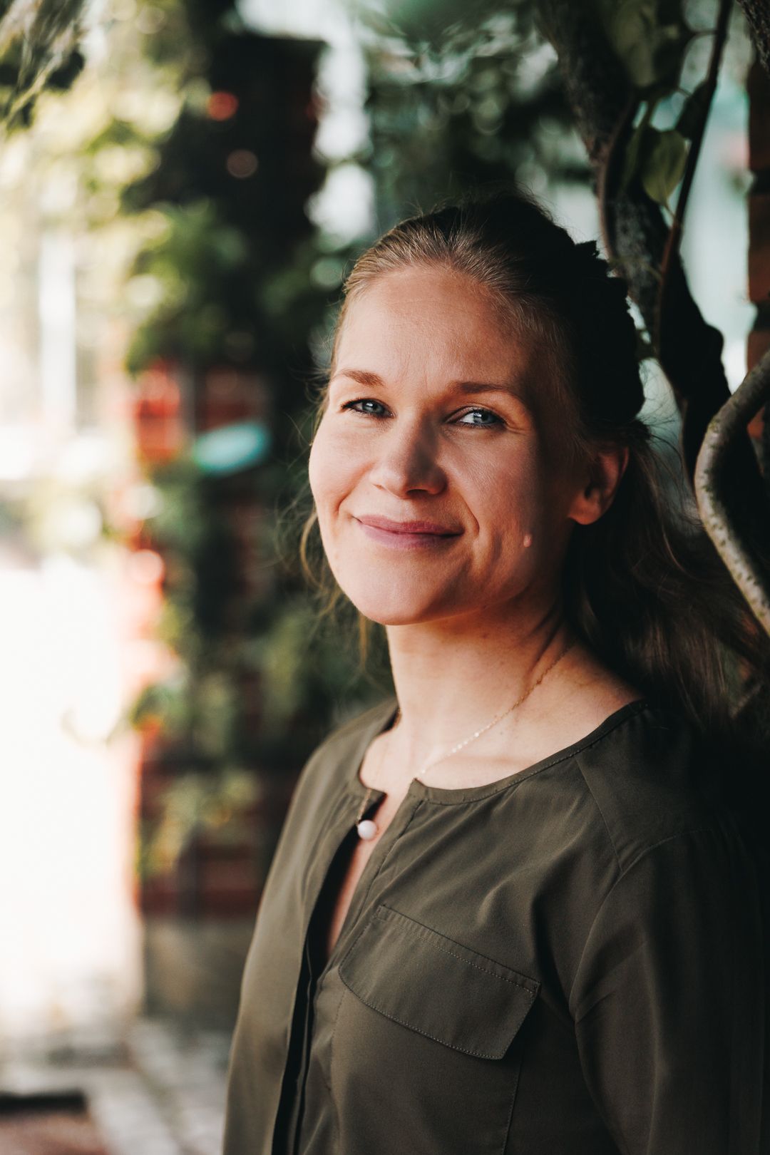 Smiling middle-aged woman with pulled-back hair wearing olive green top, posing outdoors near lush green plants.