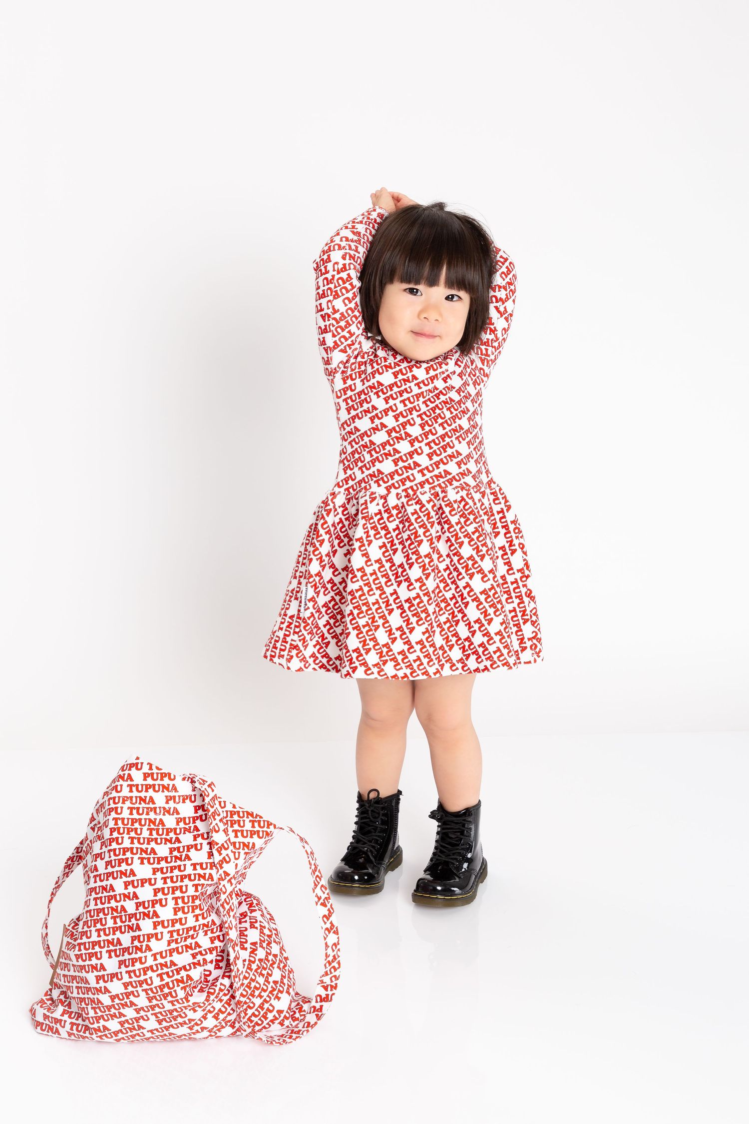 Toddler girl in red patterned dress and black boots poses playfully with arms raised on white background.