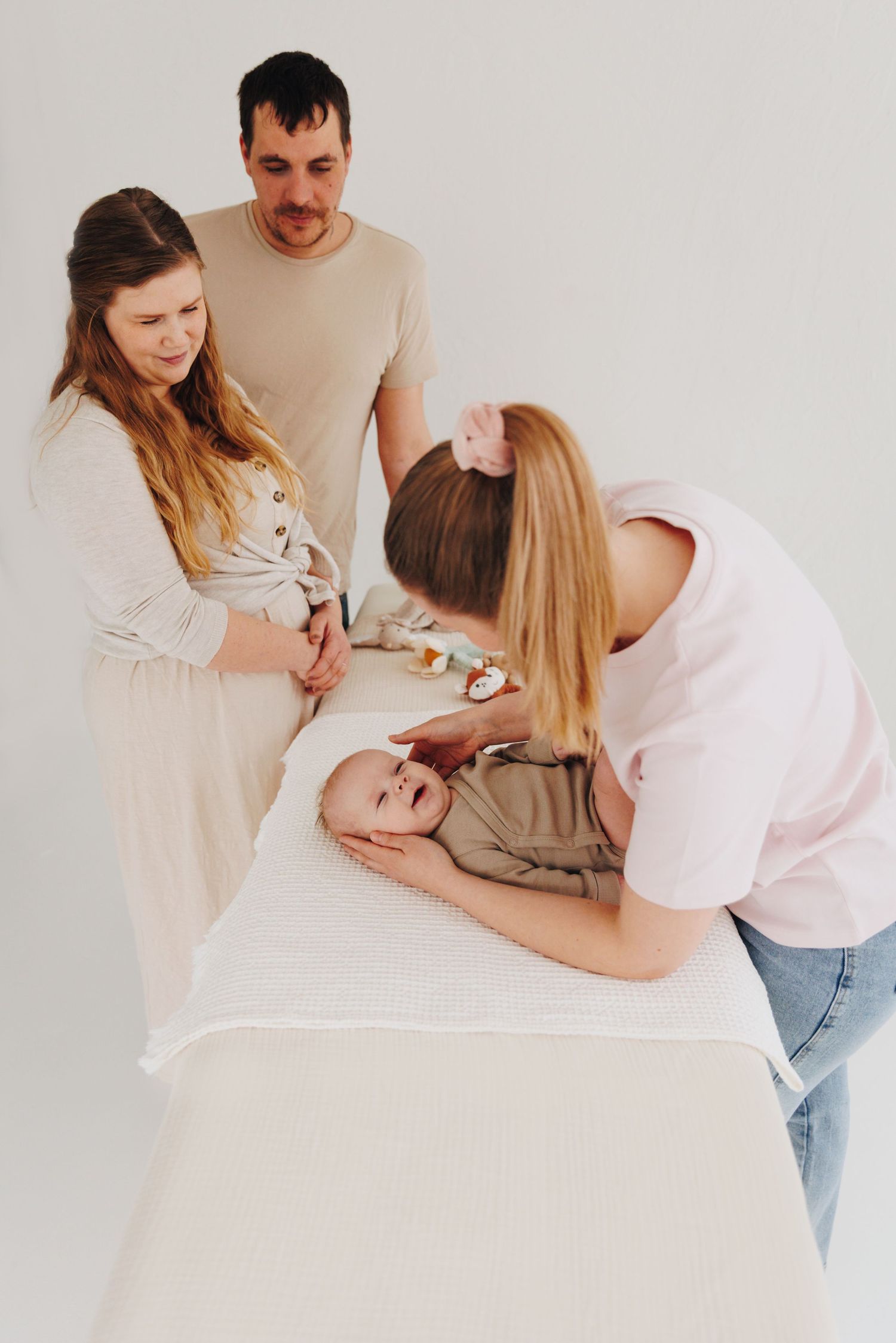 Parents and a caregiver tenderly attend to a smiling baby lying on a white table in a bright, minimalist room.