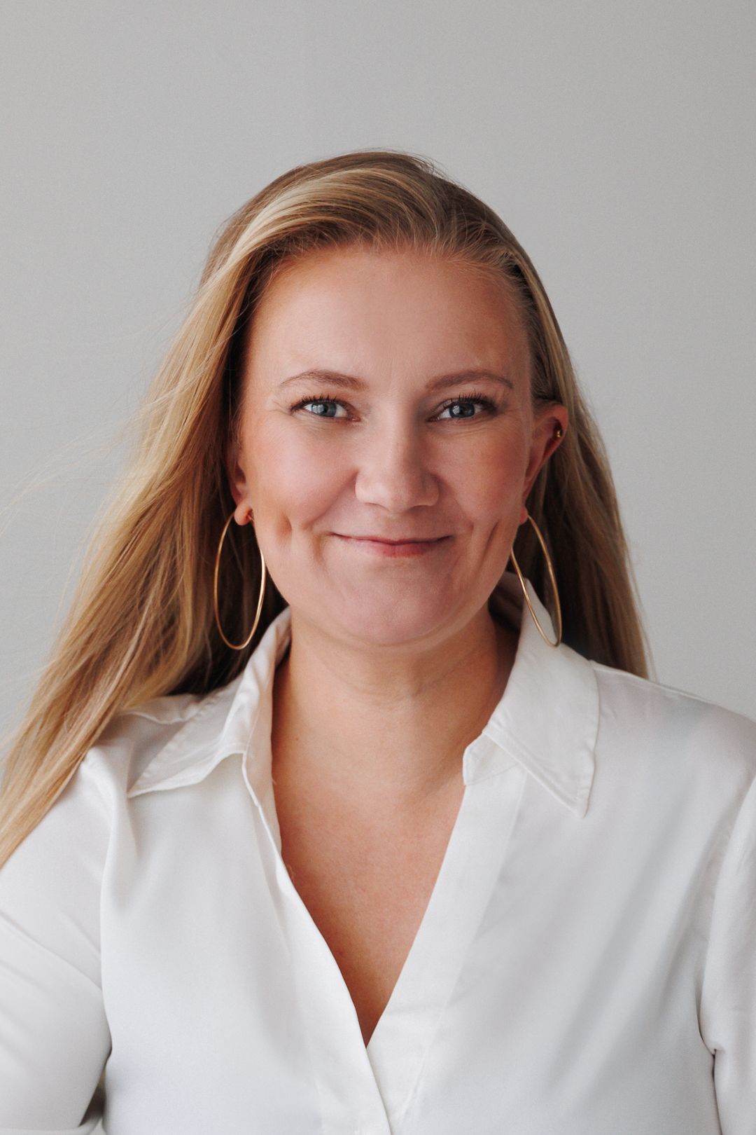 Smiling blonde woman in white blouse with gold hoop earrings poses for professional headshot photo.
