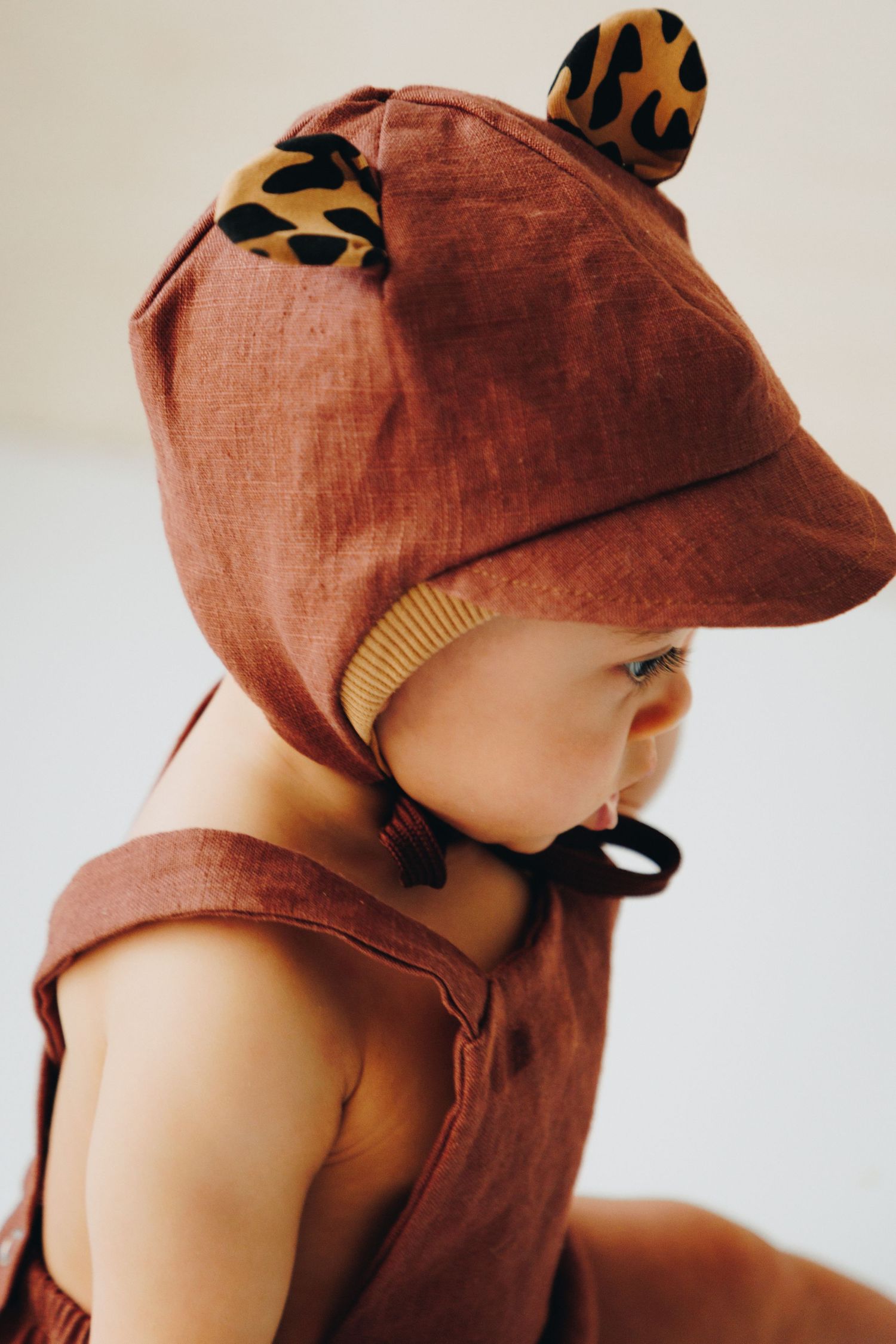 Baby wearing a rust brown linen hat with leopard print ears and matching overalls, side profile view.