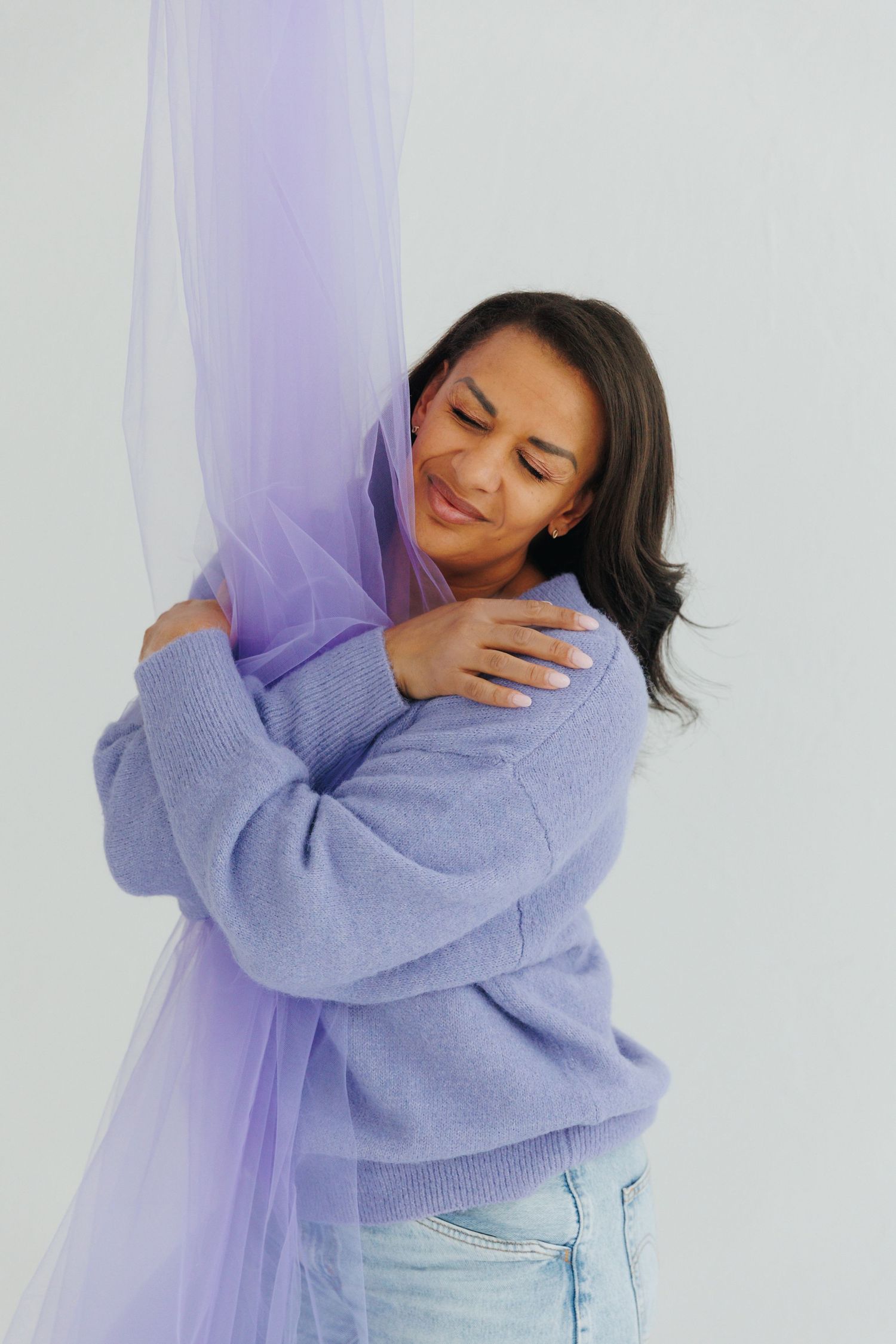 Woman in lavender sweater hugging purple tulle fabric, smiling with eyes closed against white background.