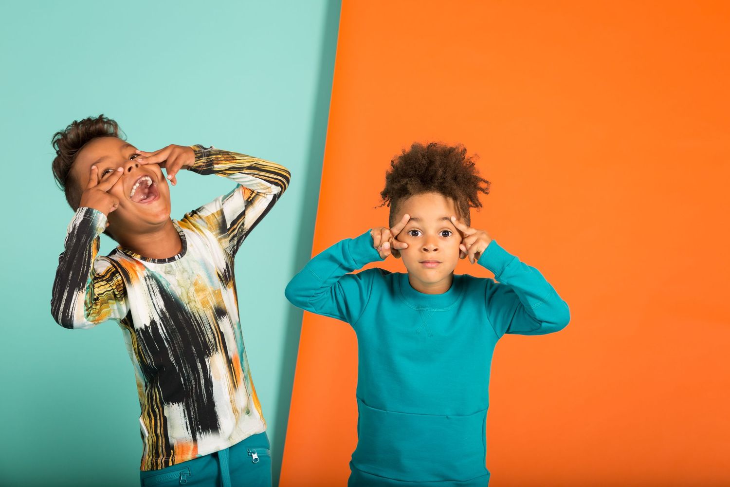 Two kids making funny faces with fingers as glasses against teal and orange background.