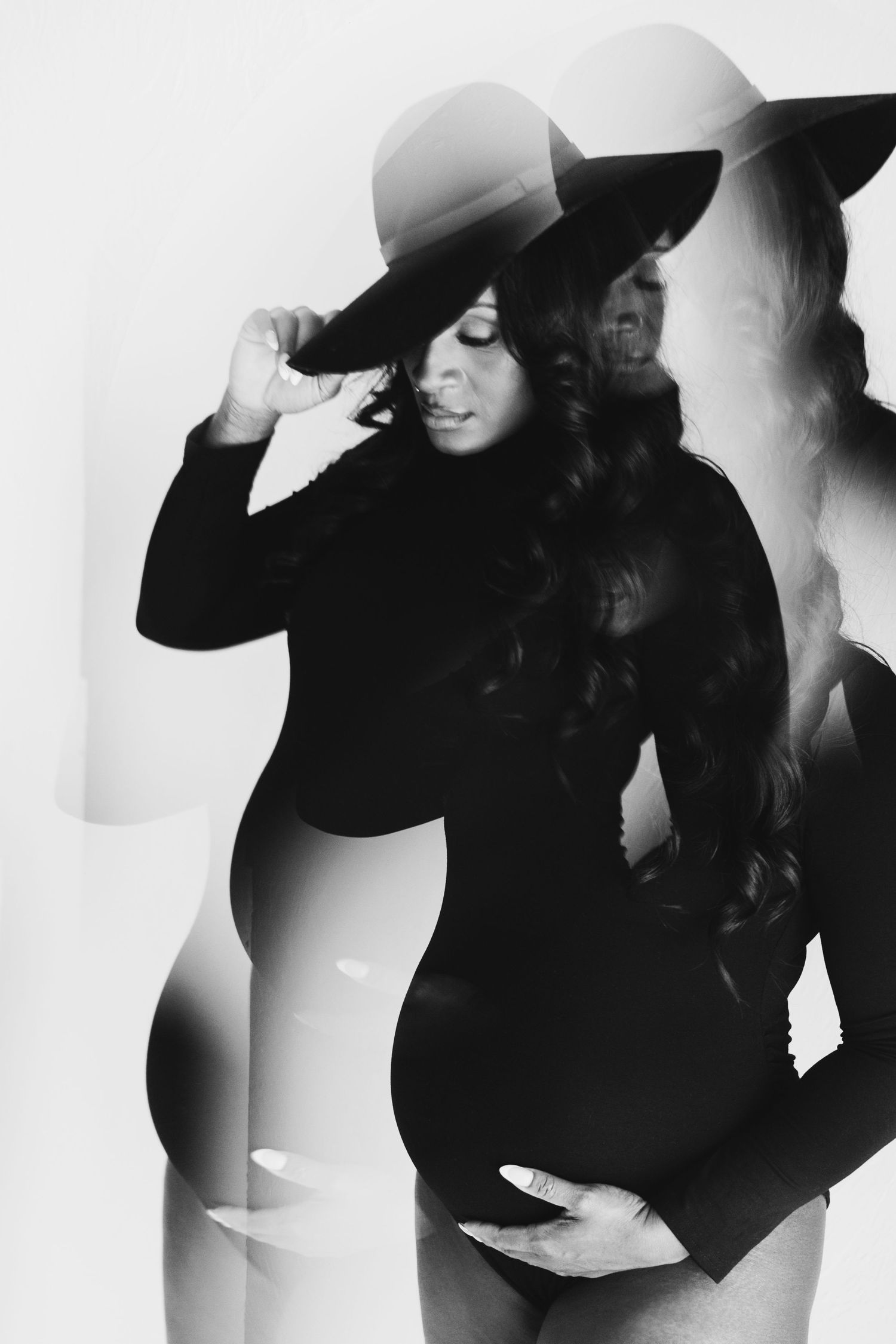 Black and white maternity photo of a pregnant woman in a black bodysuit and wide-brim hat with a double exposure effect.