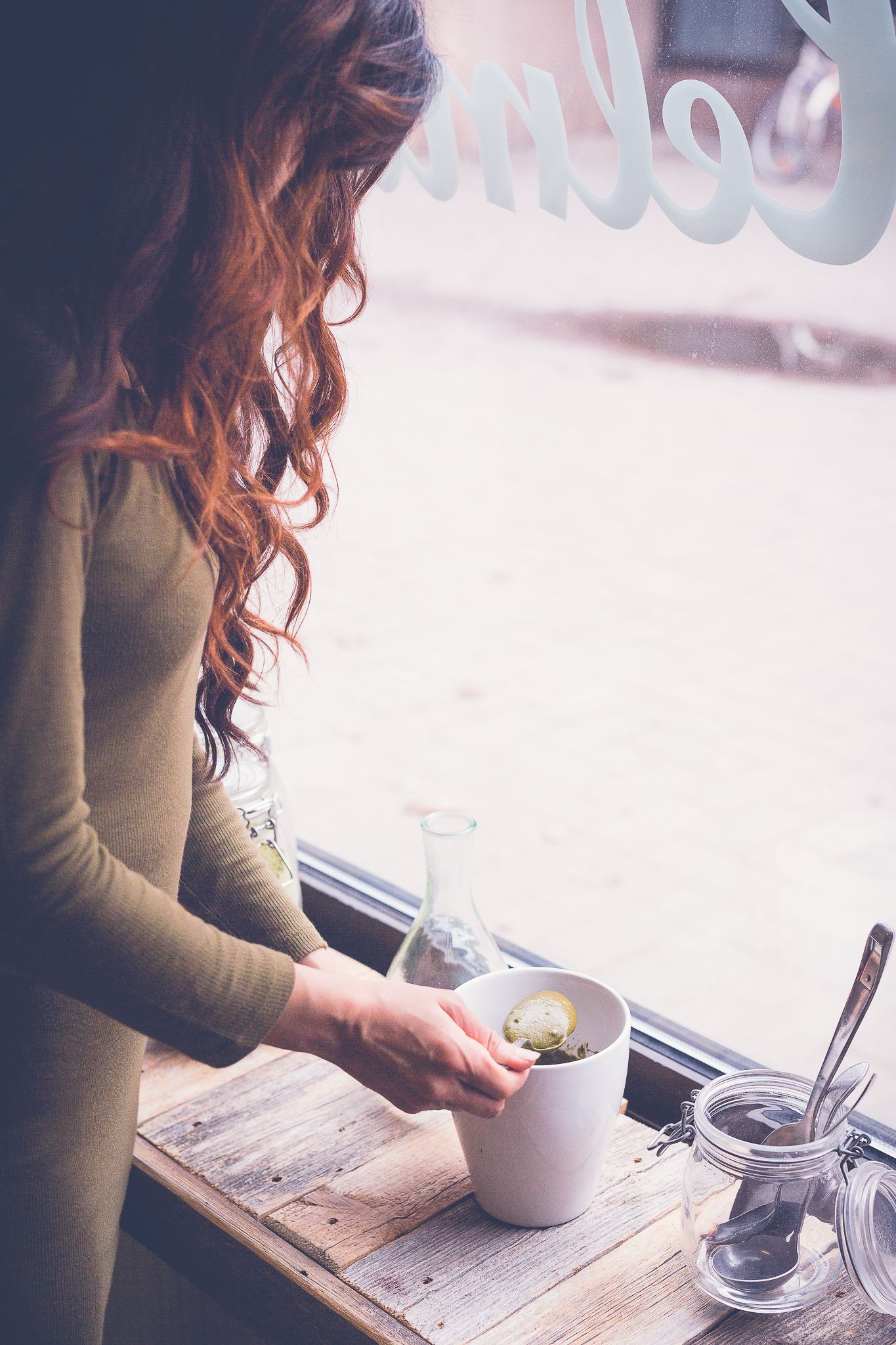 A redhead woman in an olive dress prepares a hot drink in a white mug at a rustic wooden cafe window table.