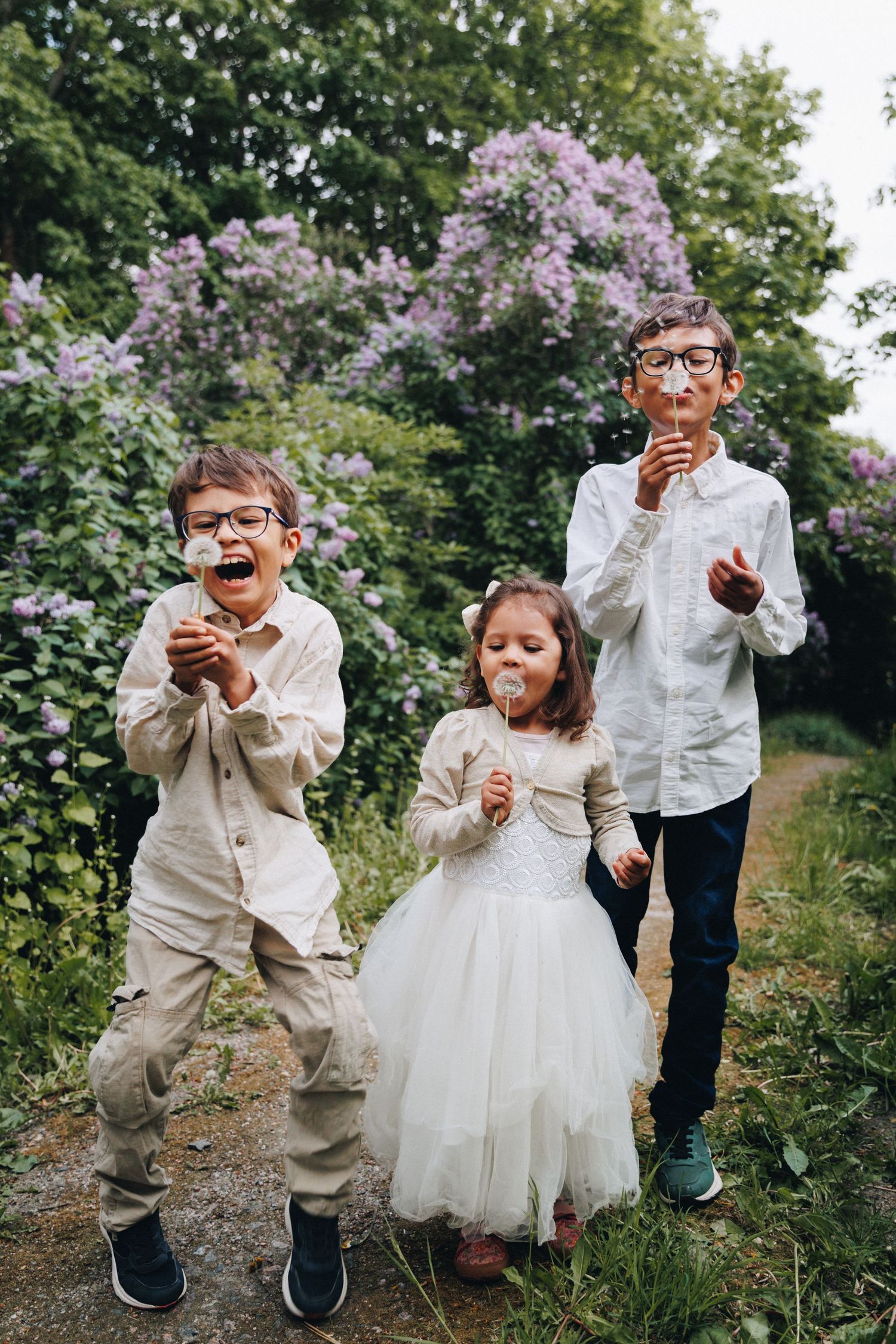 Three children blowing dandelions in a garden with purple lilac bushes, two boys wearing glasses and a girl in a white tulle dress.