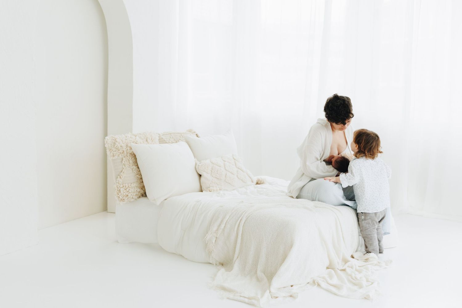 A mother sits on a white bed with her young child in a bright, minimalist white bedroom in studio at Helsinki