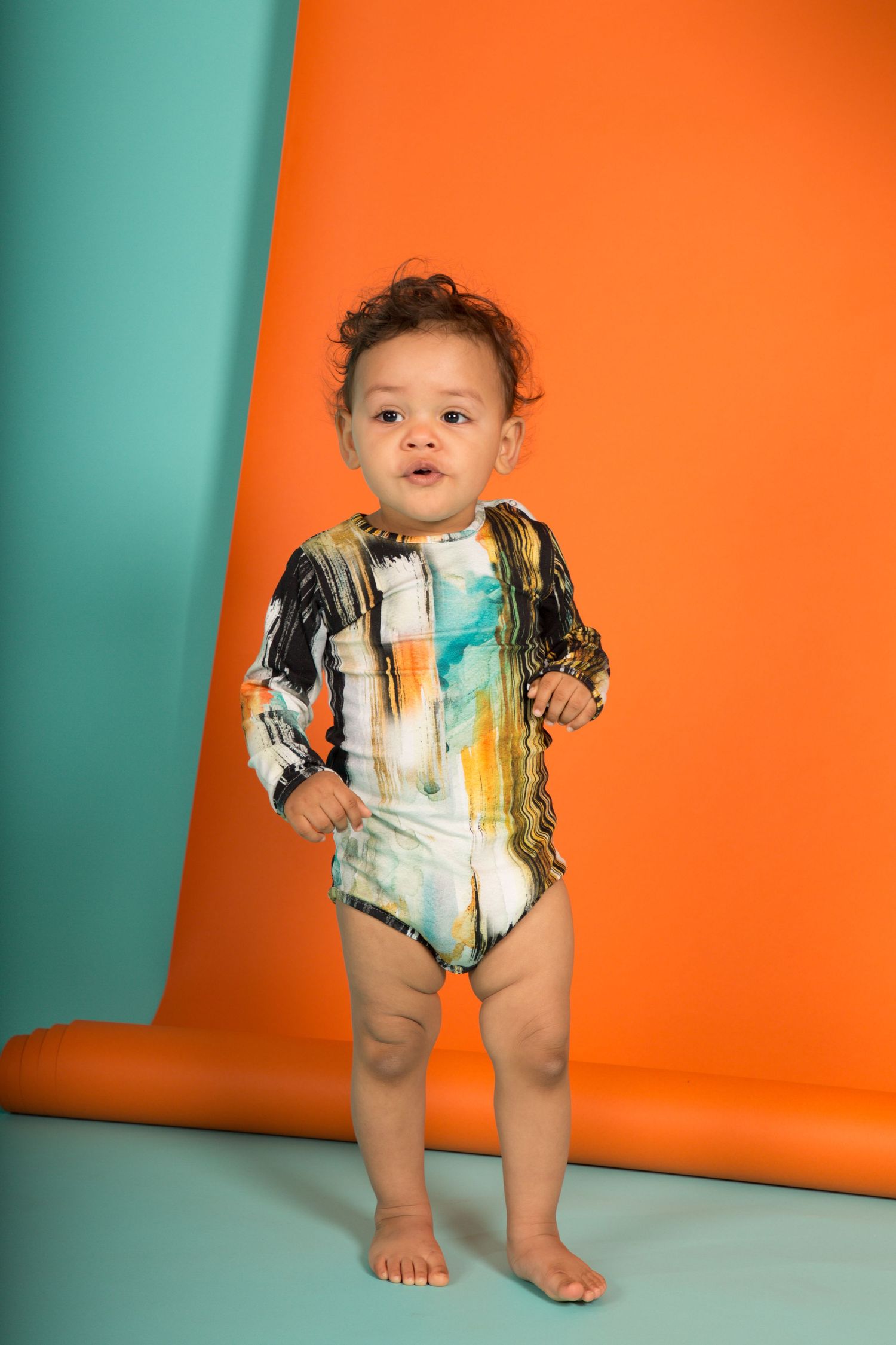 Baby stands and poses in colorful bodysuit against vibrant orange and teal background for portrait session.