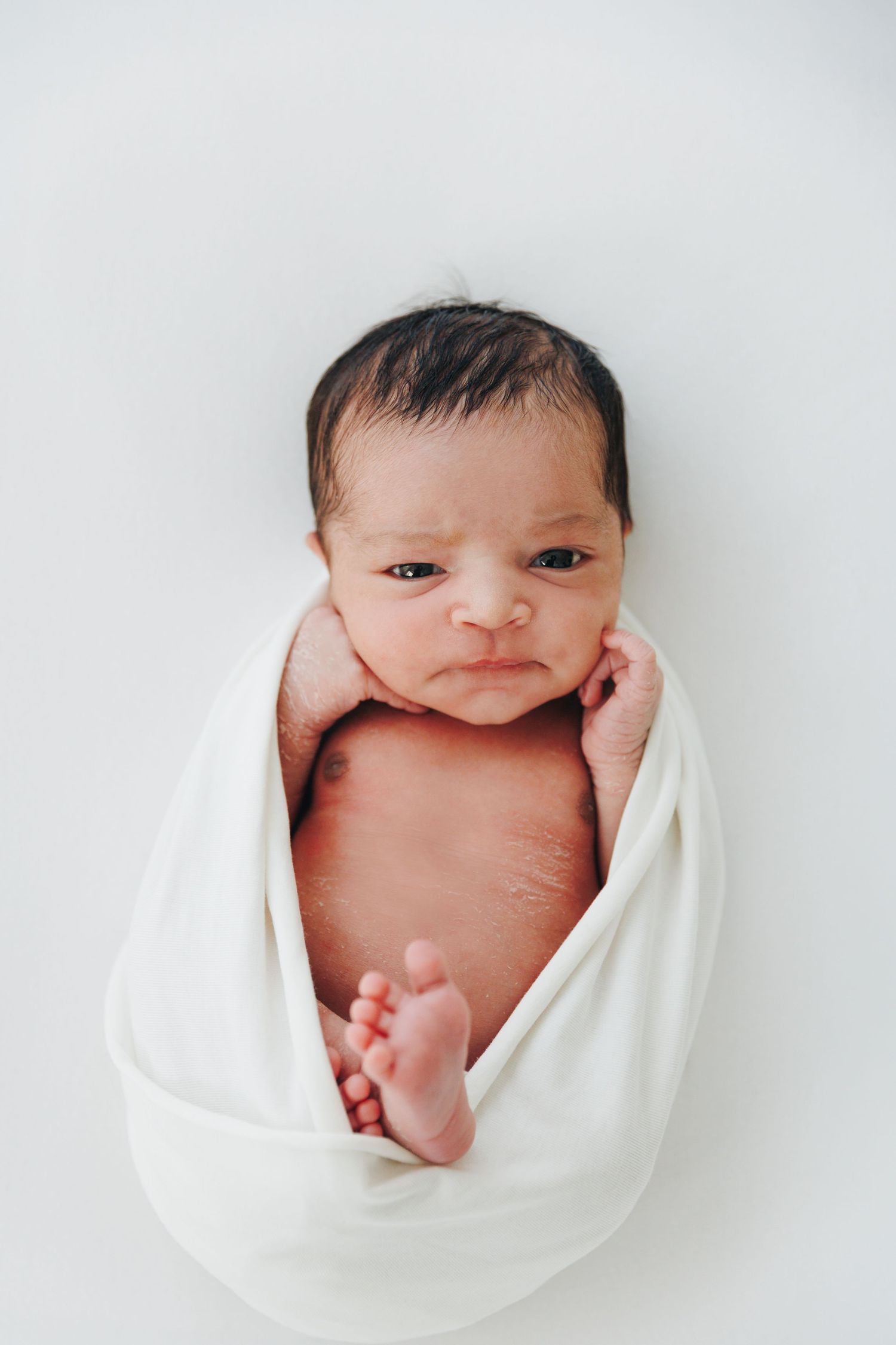 Newborn baby swaddled in white wrap lying on white background with dark hair and alert expression.