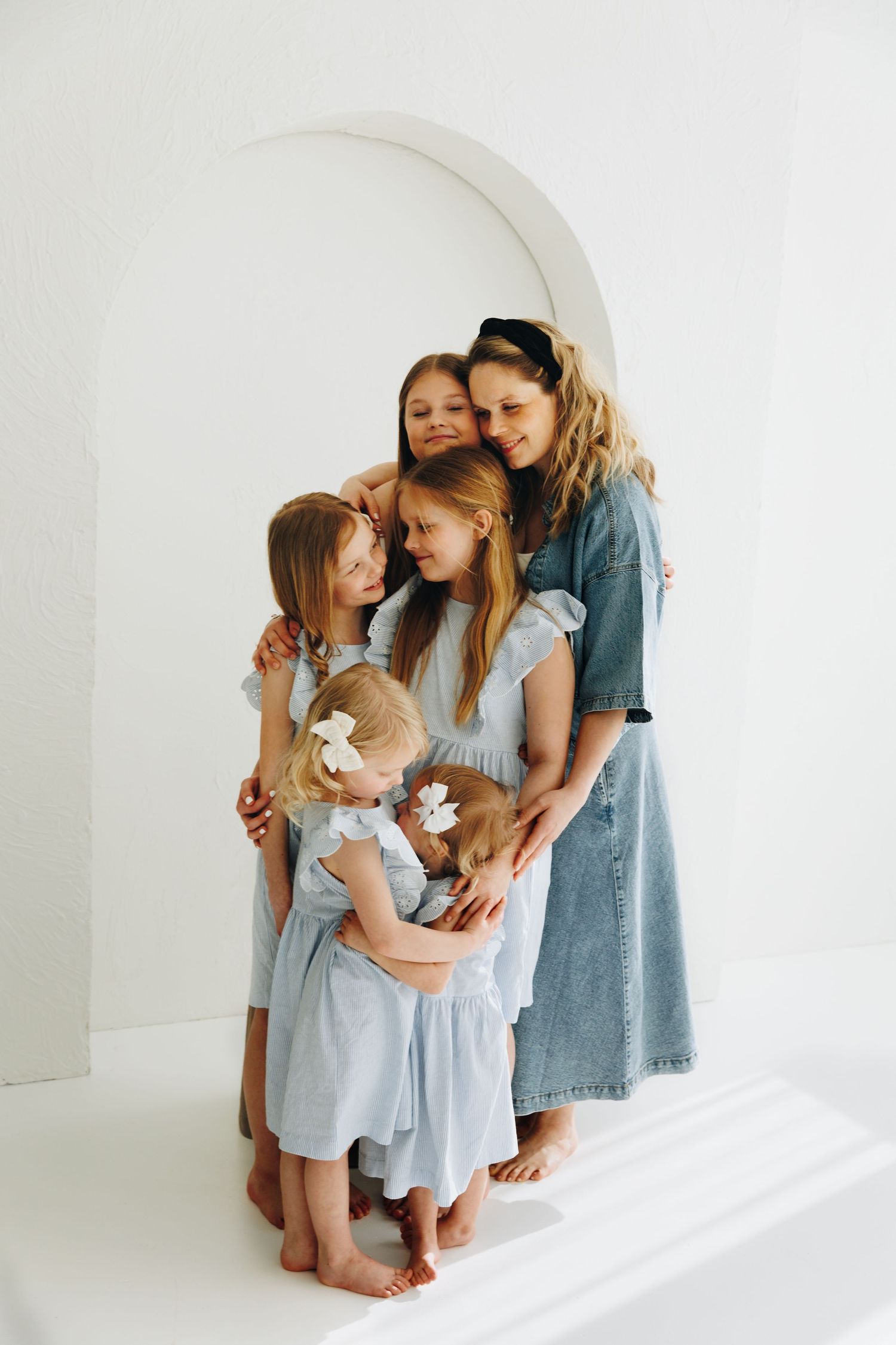 A woman in a denim dress hugs four young girls in light blue dresses against a white arched backdrop.