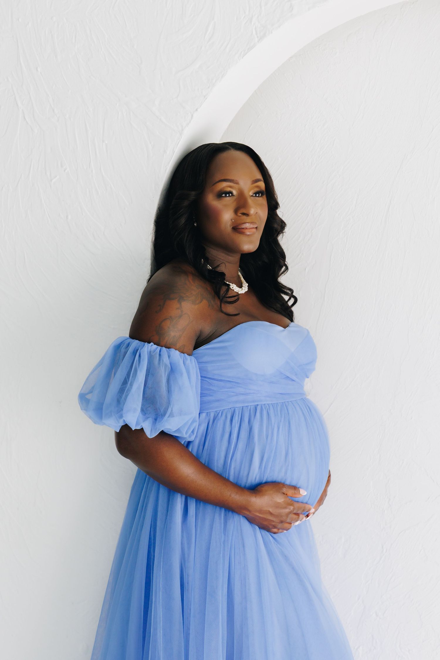 Pregnant woman in blue tulle off-shoulder gown cradling baby bump, posing against white wall.
