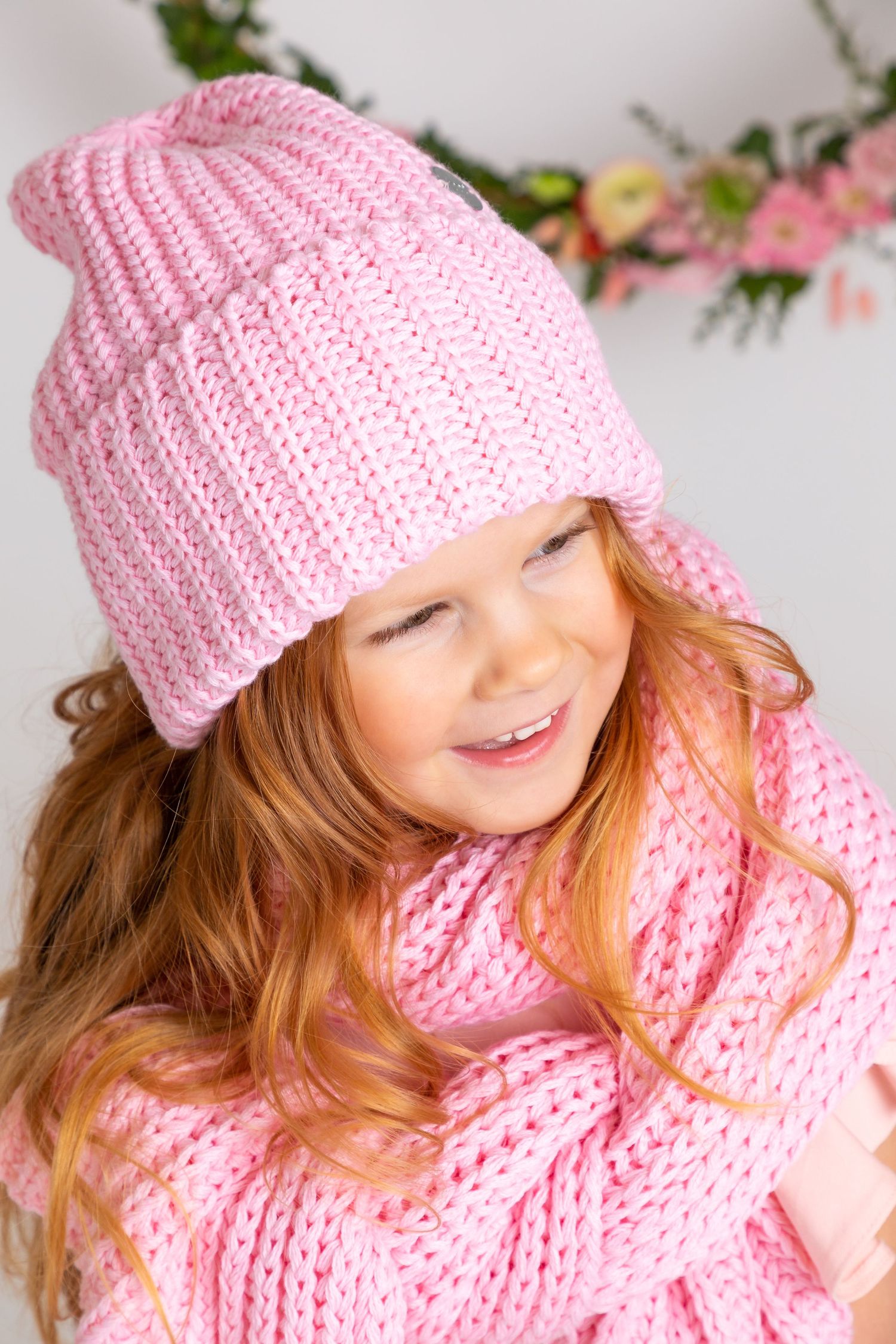 Happy young girl with red hair wearing a cozy pink knit hat and scarf, smiling joyfully in a winter outfit.
