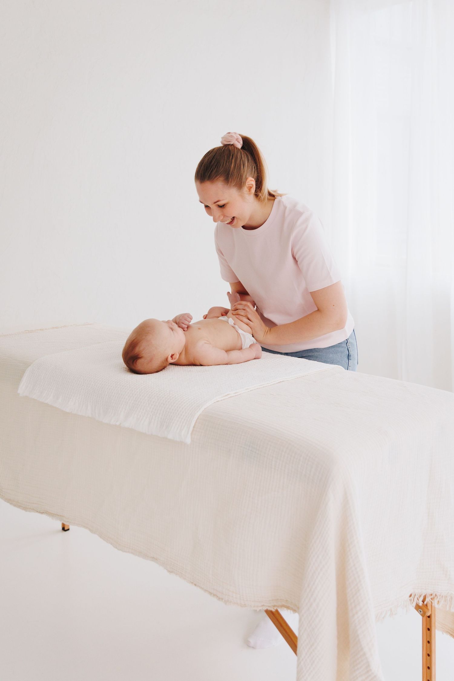 A young woman gently massages a baby lying on a white massage table in a bright, clean therapy room.