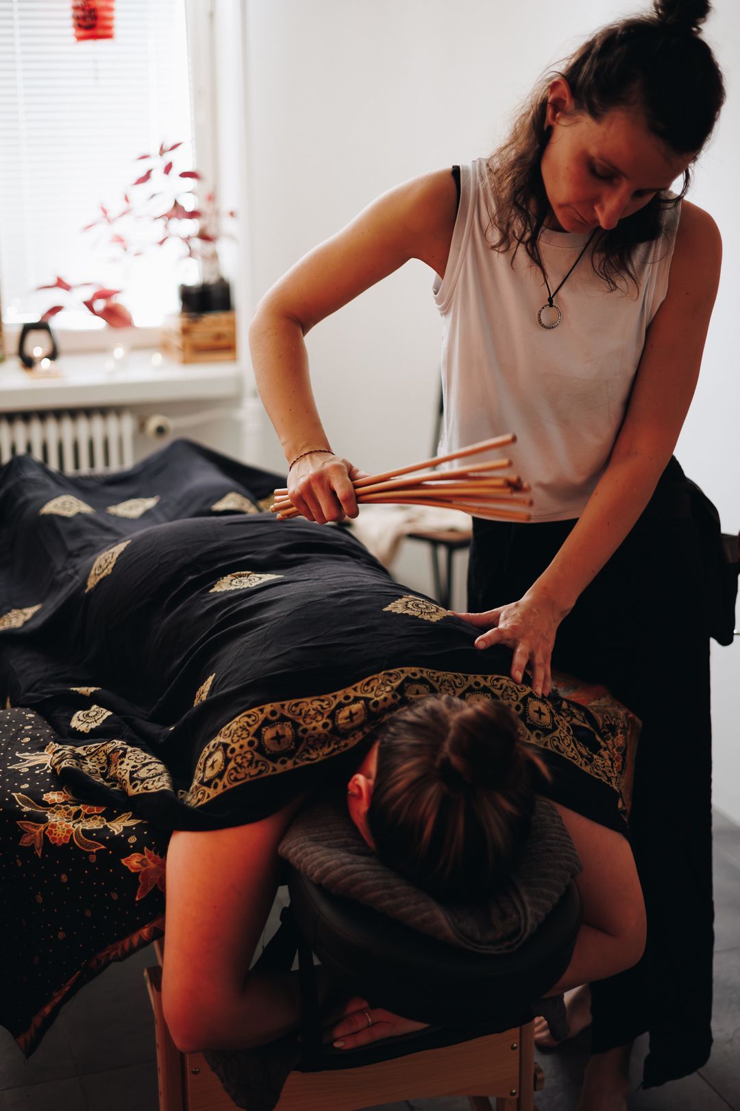 Massage therapist performing a bamboo stick massage on a client lying face down on a decorated massage table.