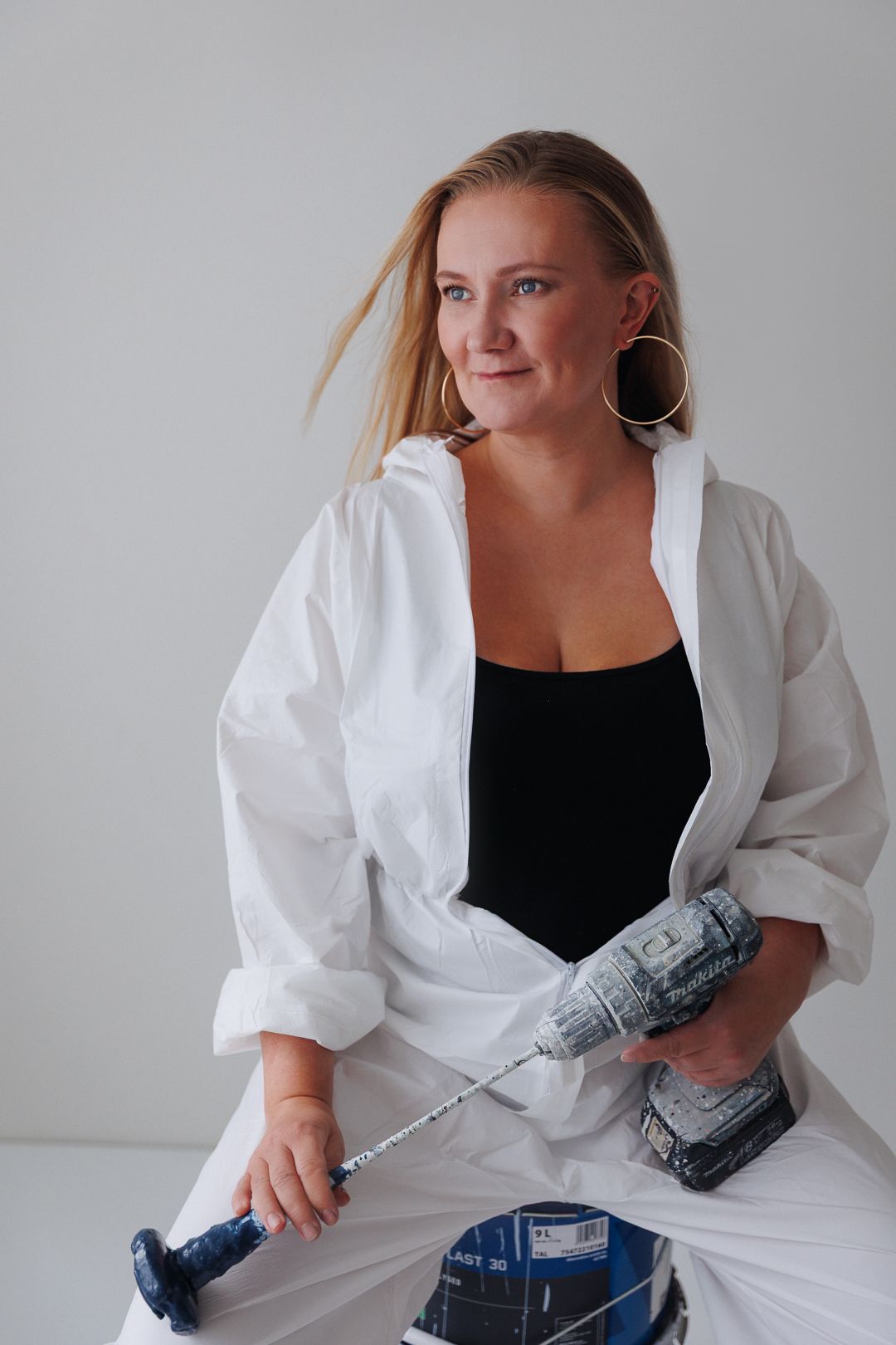 Blonde woman in white overalls holding a power drill, sitting against a light background.