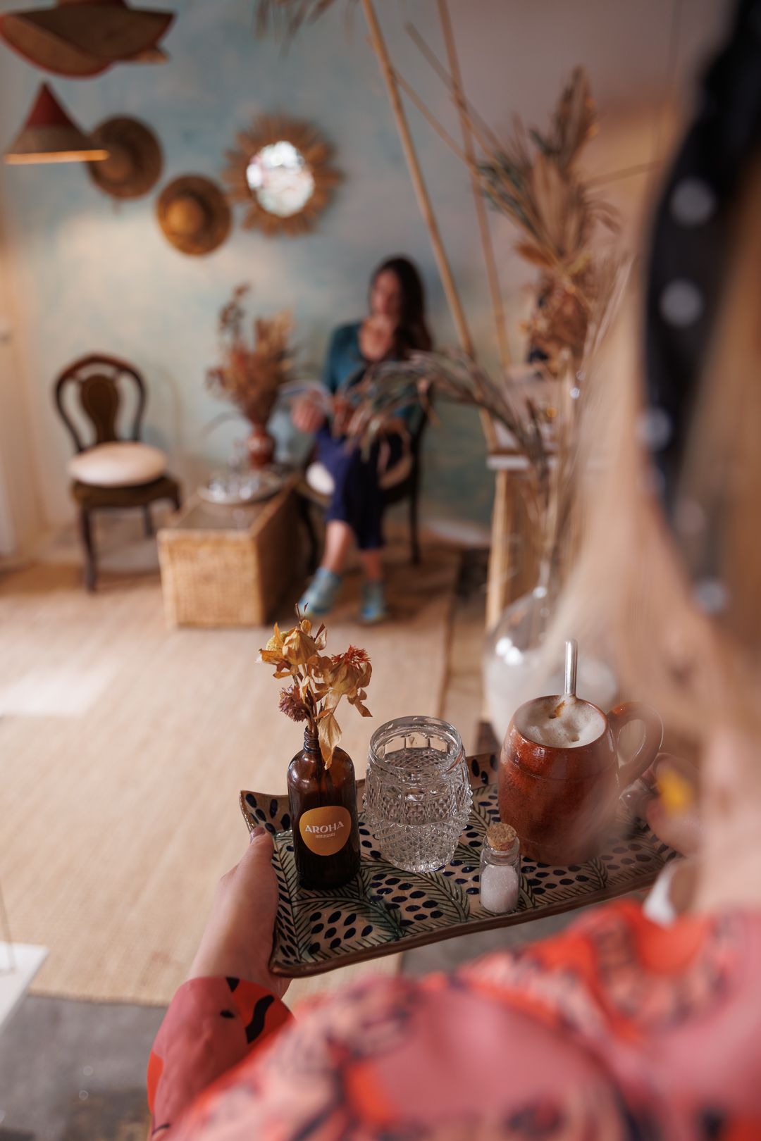 Person holding a decorative tray with drinks and dried flowers in a cozy bohemian-style cafe interior.
