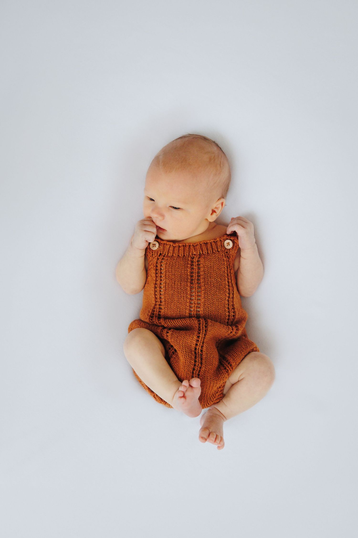 Newborn baby in rust knit romper lying on white background, looking sideways with hands near face.