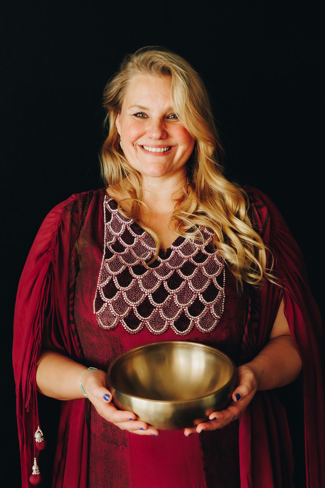 Smiling blonde woman in red dress holding a golden singing bowl against black background.