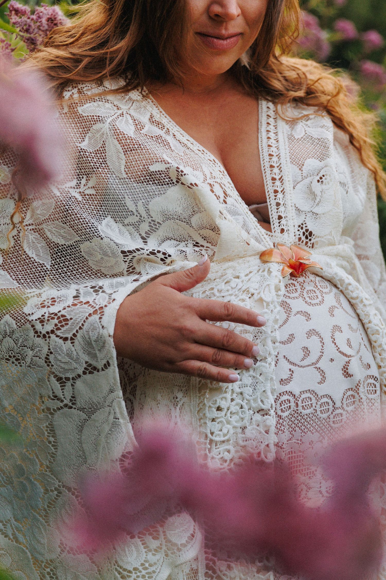 Pregnant woman in white lace maternity gown cradling her baby bump outdoors among pink blooming flowers.