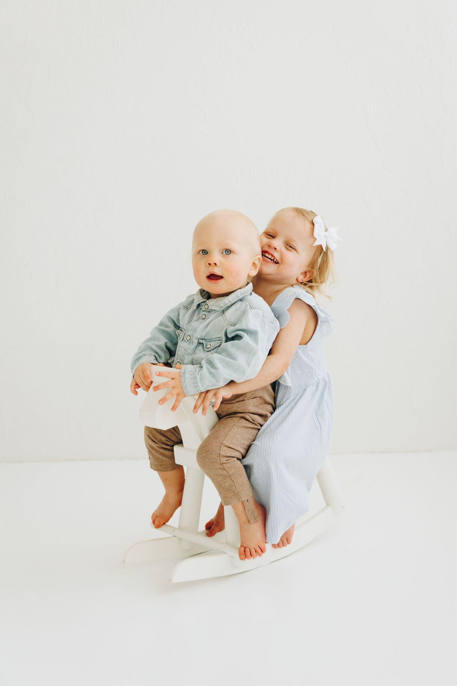 Two young siblings pose together, a baby boy in denim shirt on a rocking horse with his smiling older sister in a blue dress.