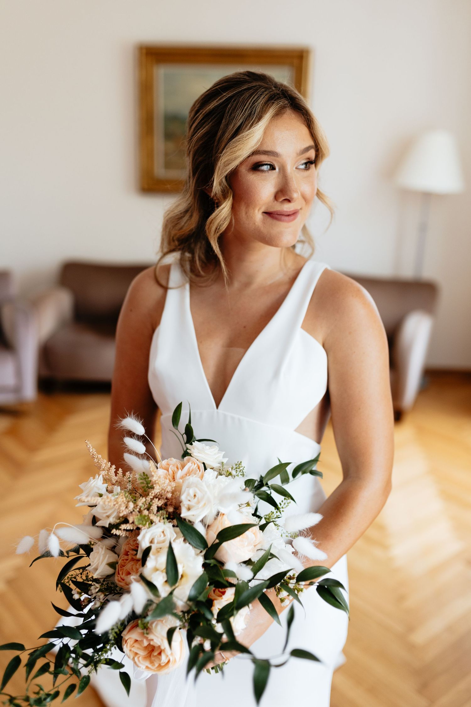 Elegant bride in white V-neck dress holding a soft pink and white floral bouquet in an indoor setting.