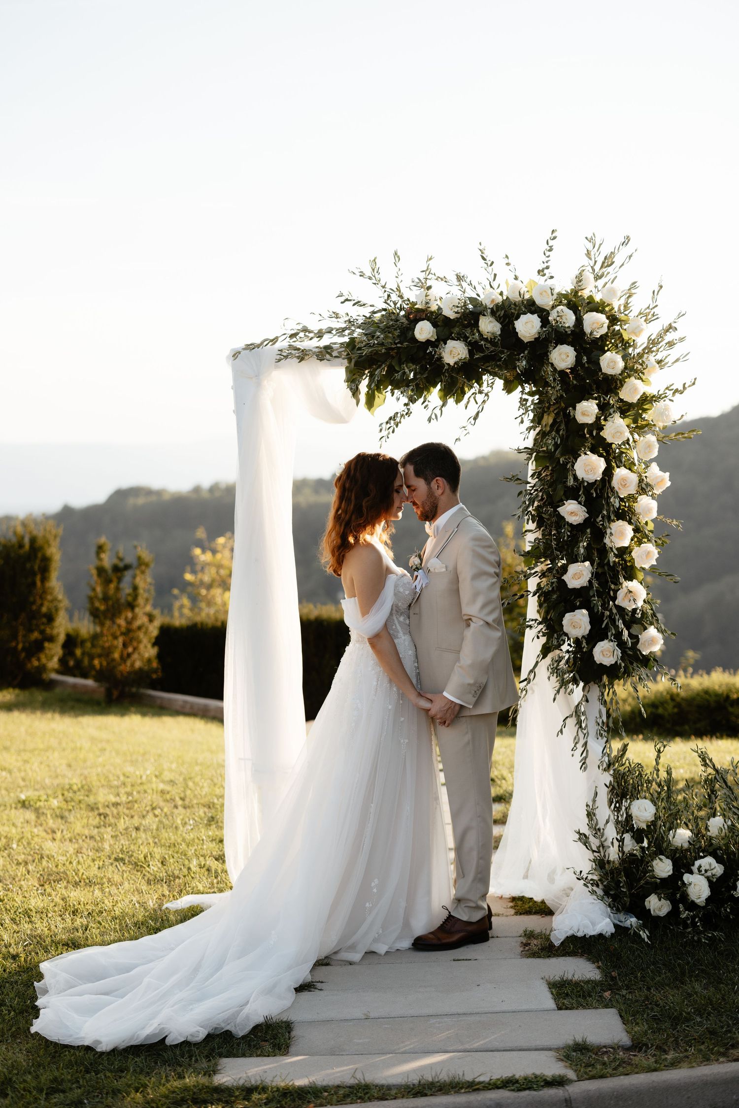 Elegant outdoor wedding ceremony setup with white floral arch and flowing drapery against mountain backdrop.