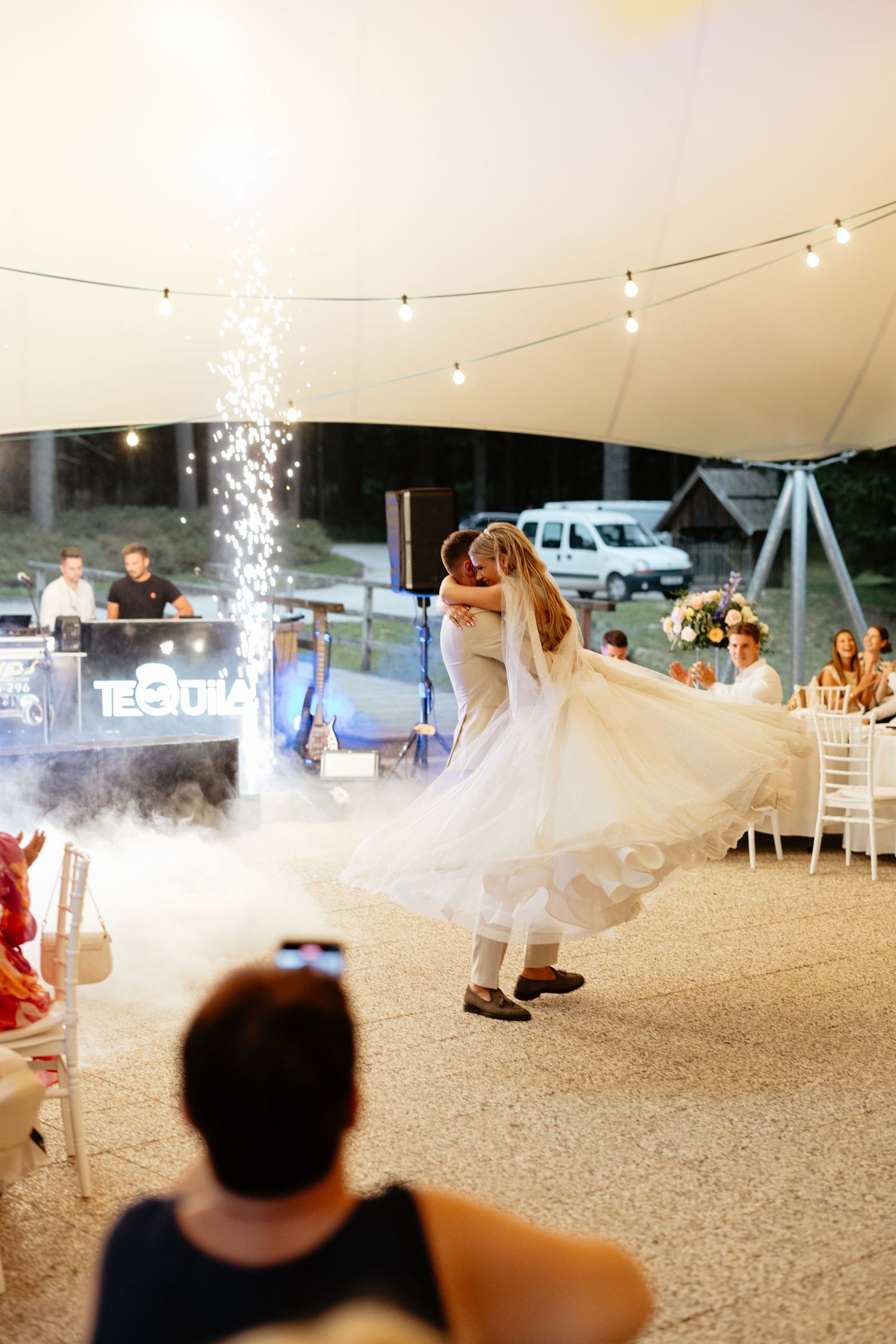 Wedding reception guests dance under string lights and sparklers in a white tent celebration.