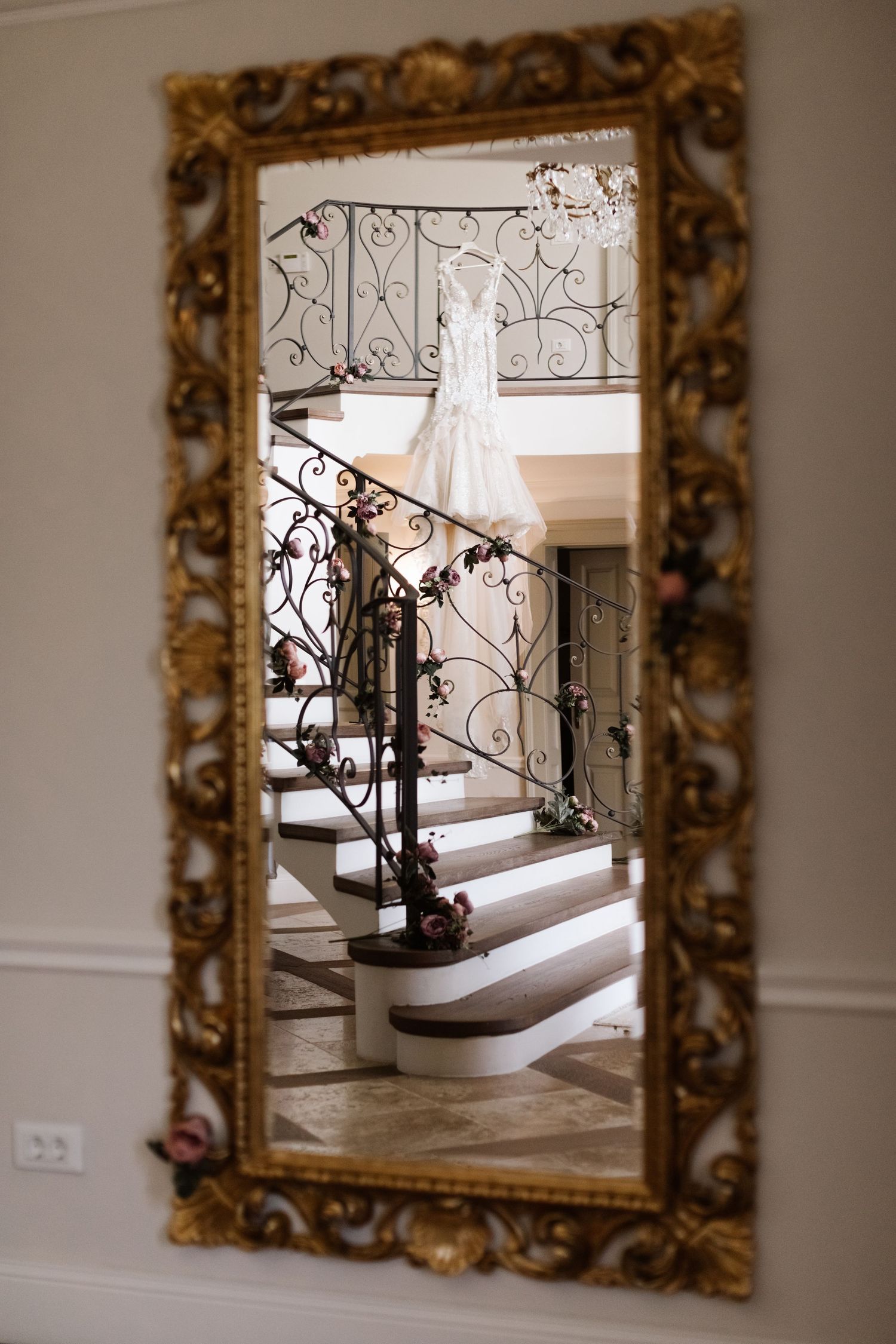 Ornate gold-framed mirror reflects elegant staircase with wrought iron railings in a luxurious interior setting.