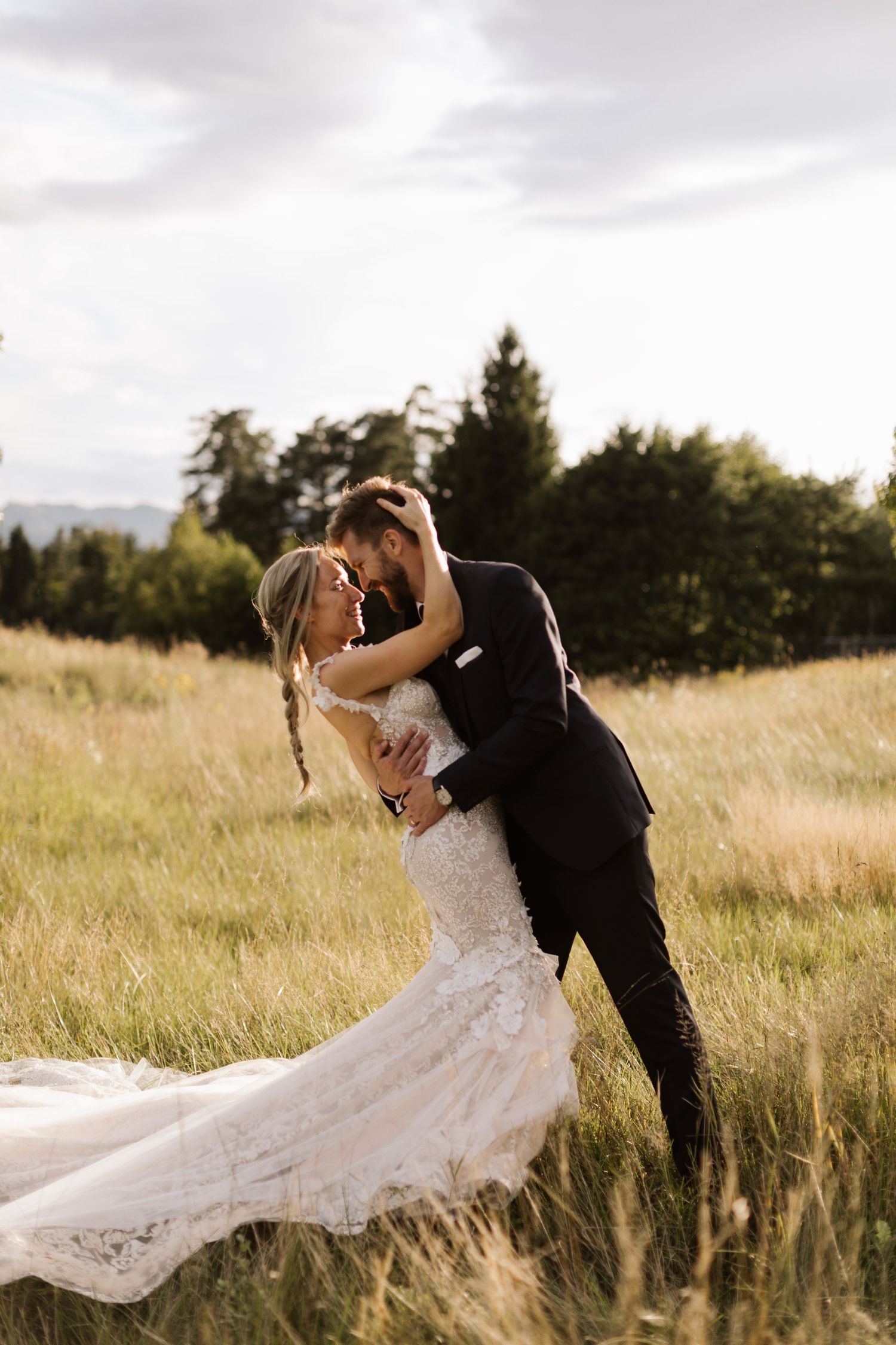 A romantic wedding couple embraces in a golden field at sunset with mountains and evergreen trees in the background.