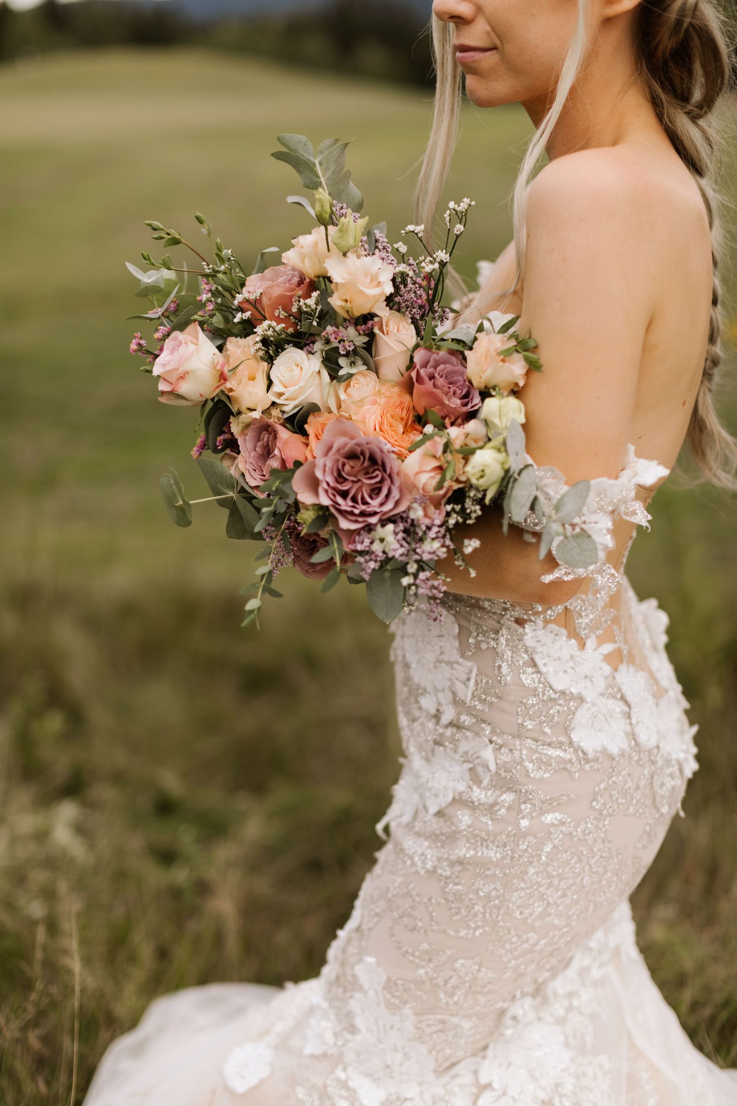 Close-up detail of a bride wearing an ornate lace wedding dress while holding a pink and mauve floral bouquet.