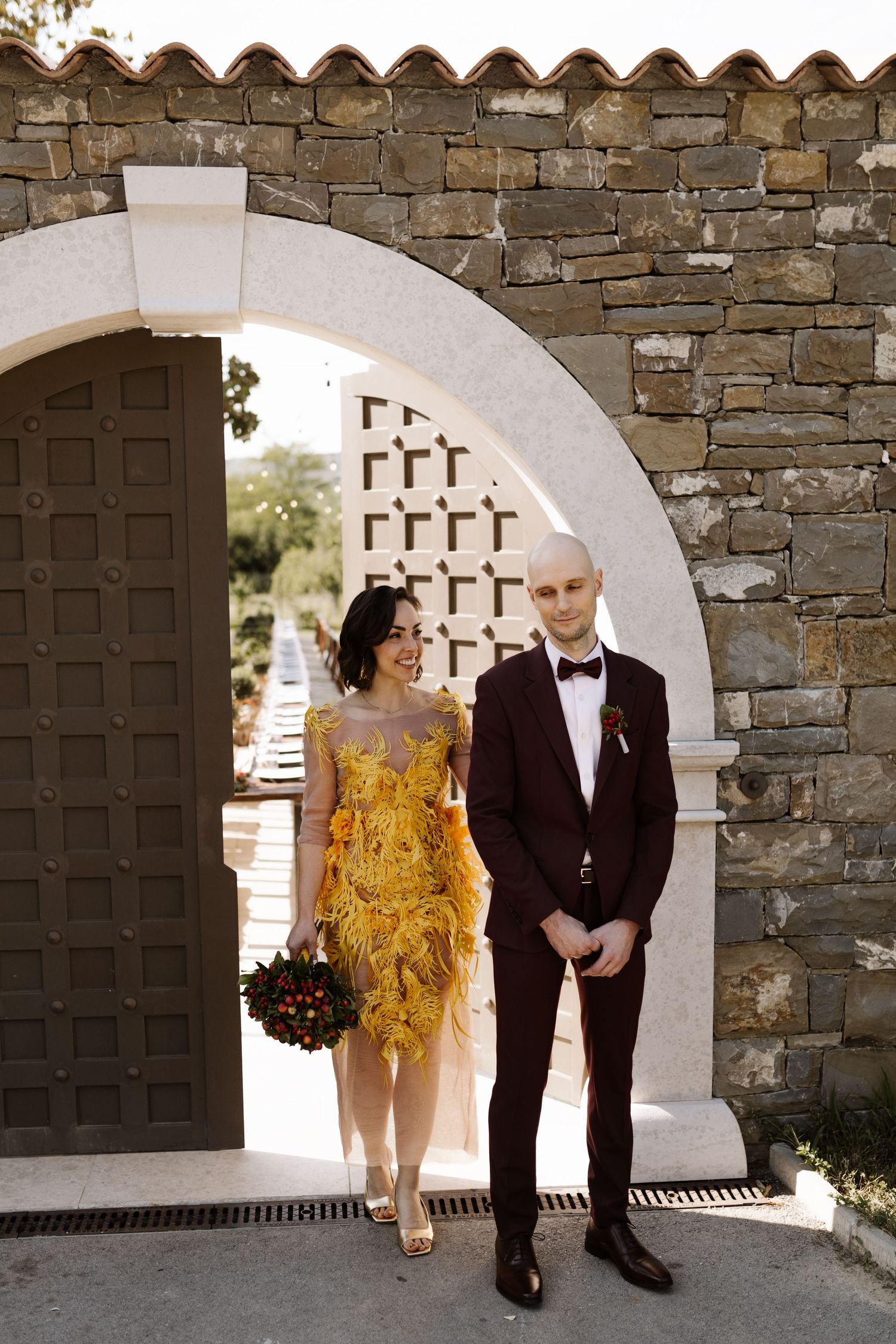A couple poses in formal attire beneath a stone archway with rustic wooden doors.