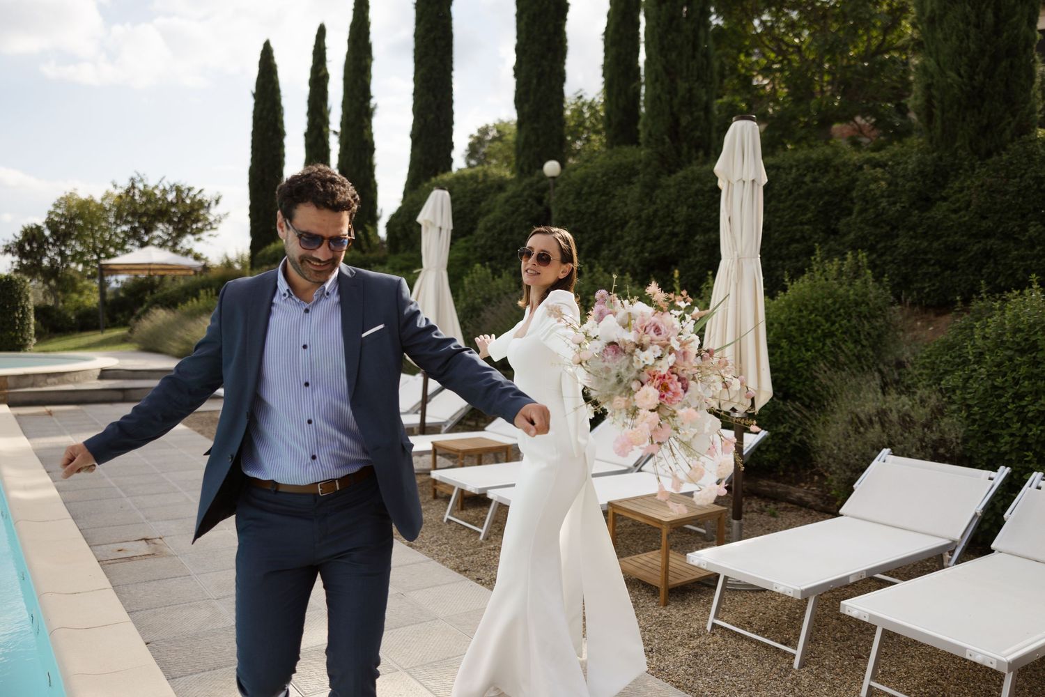 A bride and groom walk hand in hand through a Mediterranean garden setting with cypress trees and white umbrellas.