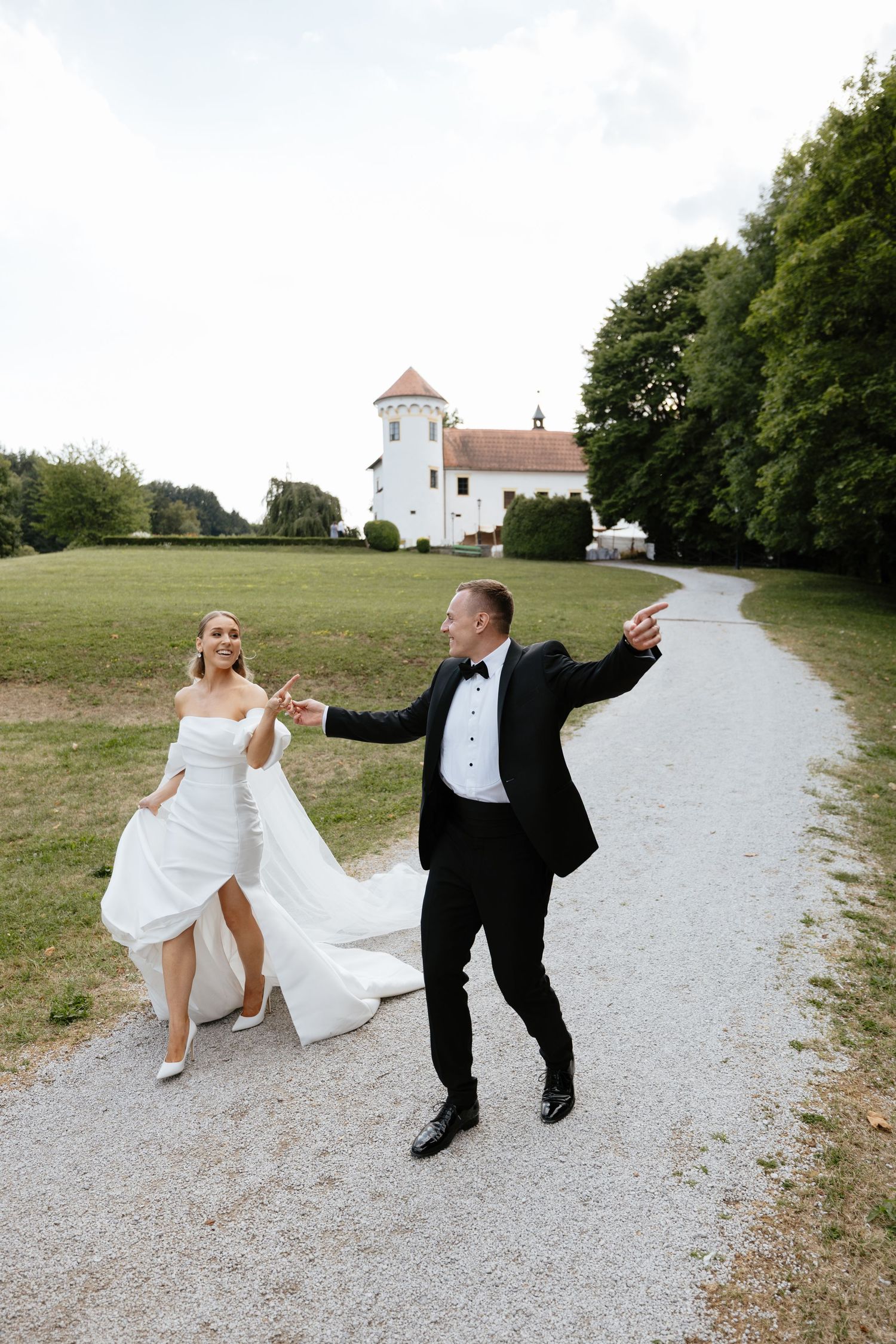 Playful wedding couple dancing on gravel path with white church visible in background.