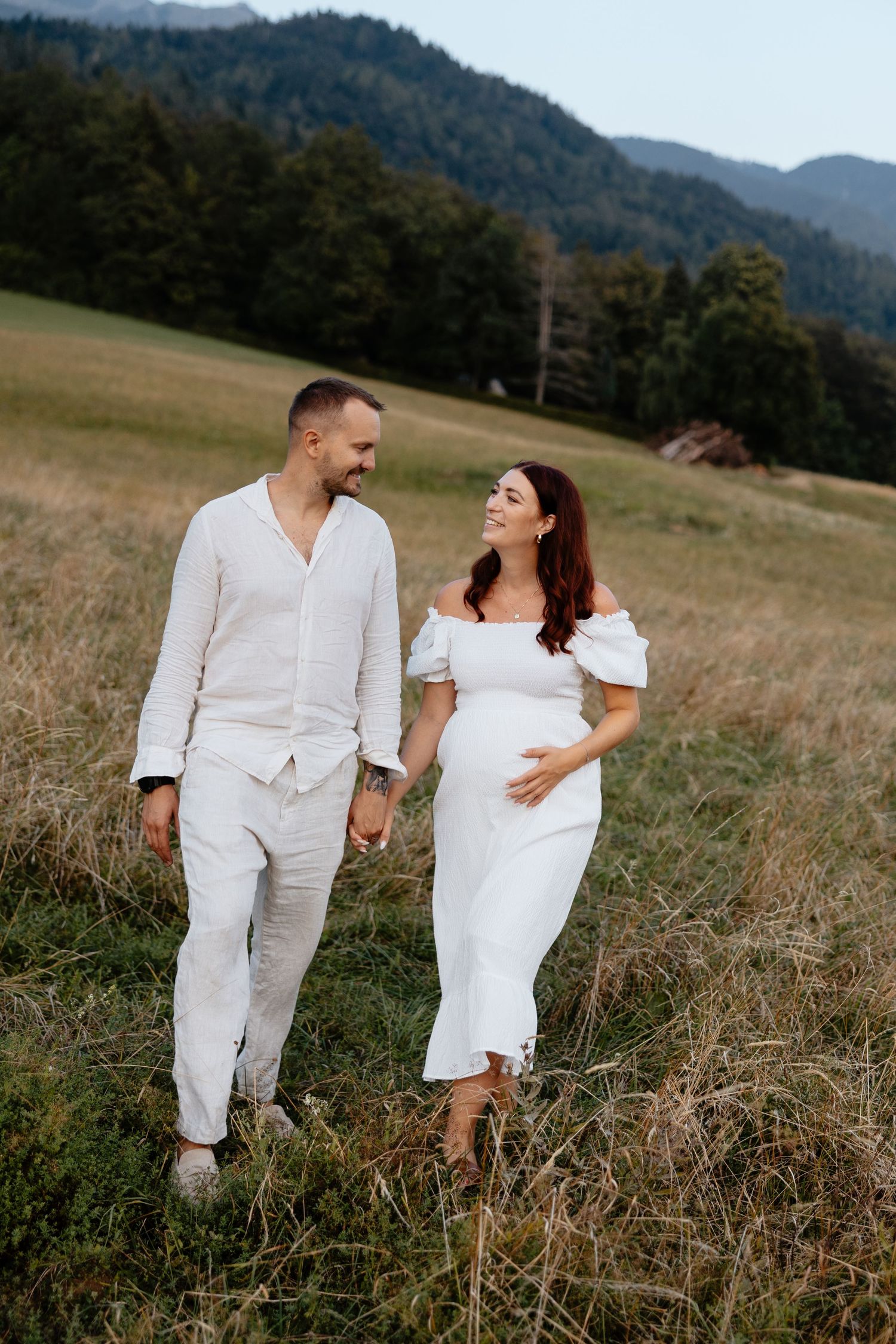 A couple in white outfits shares intimate moments in a grassy field with mountains in the background.