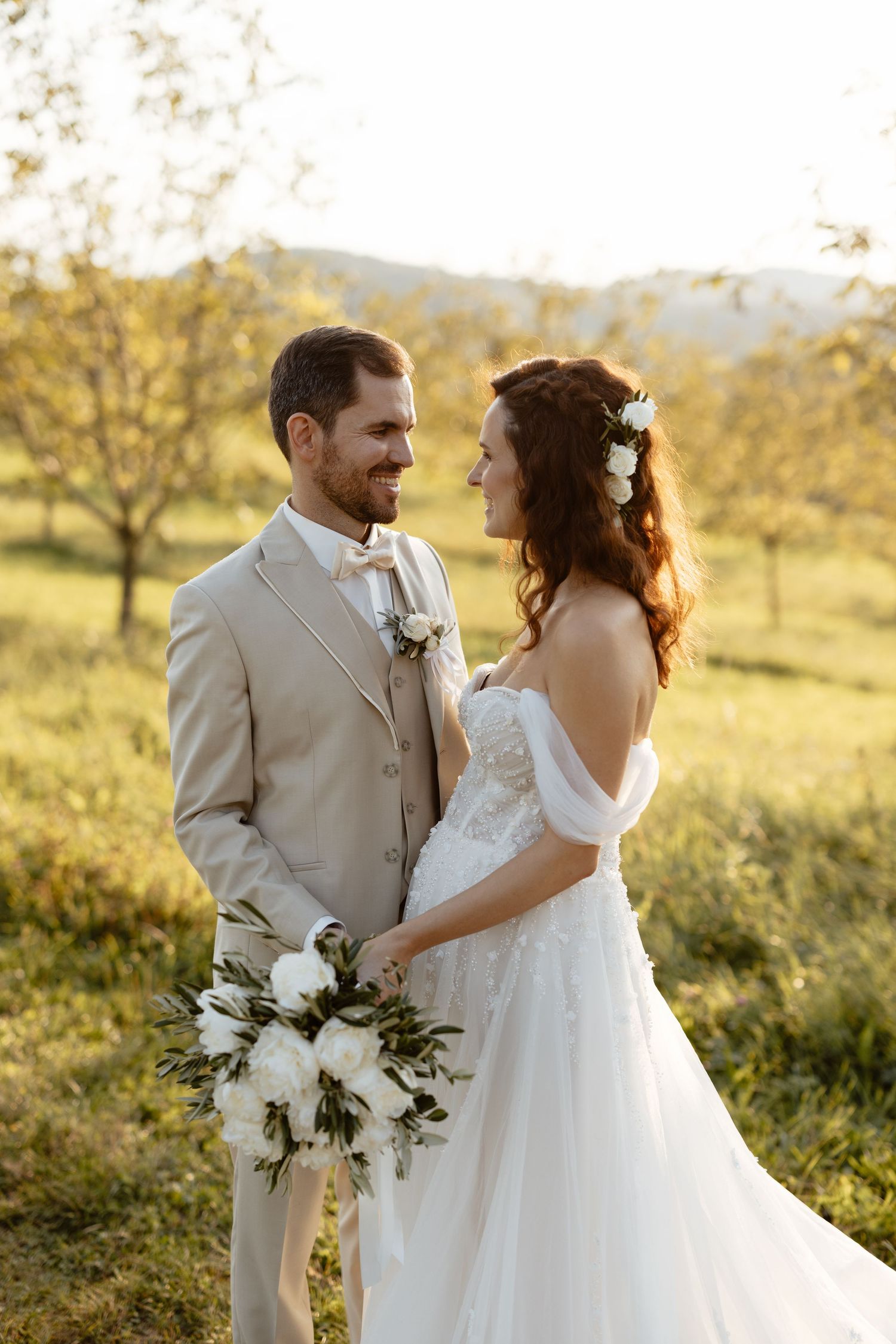 Romantic wedding portrait in golden sunlight with couple holding white bouquet in meadow setting.