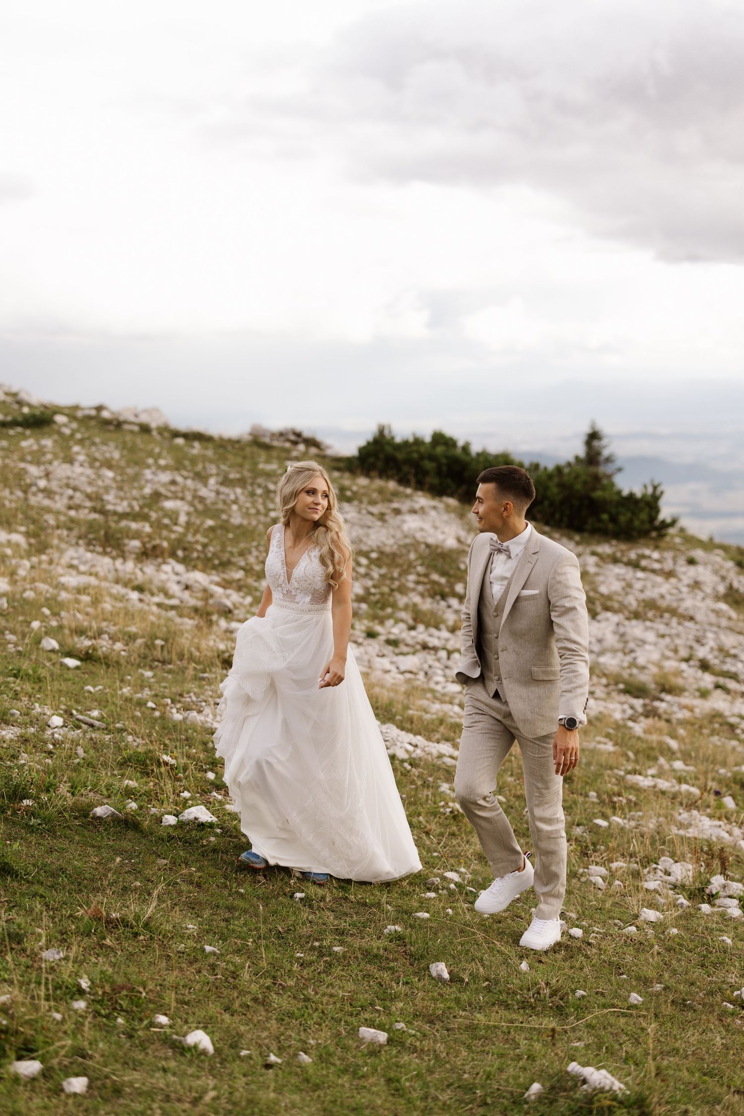 Couple in wedding attire walking together on a scenic grassy hilltop with cloudy skies.