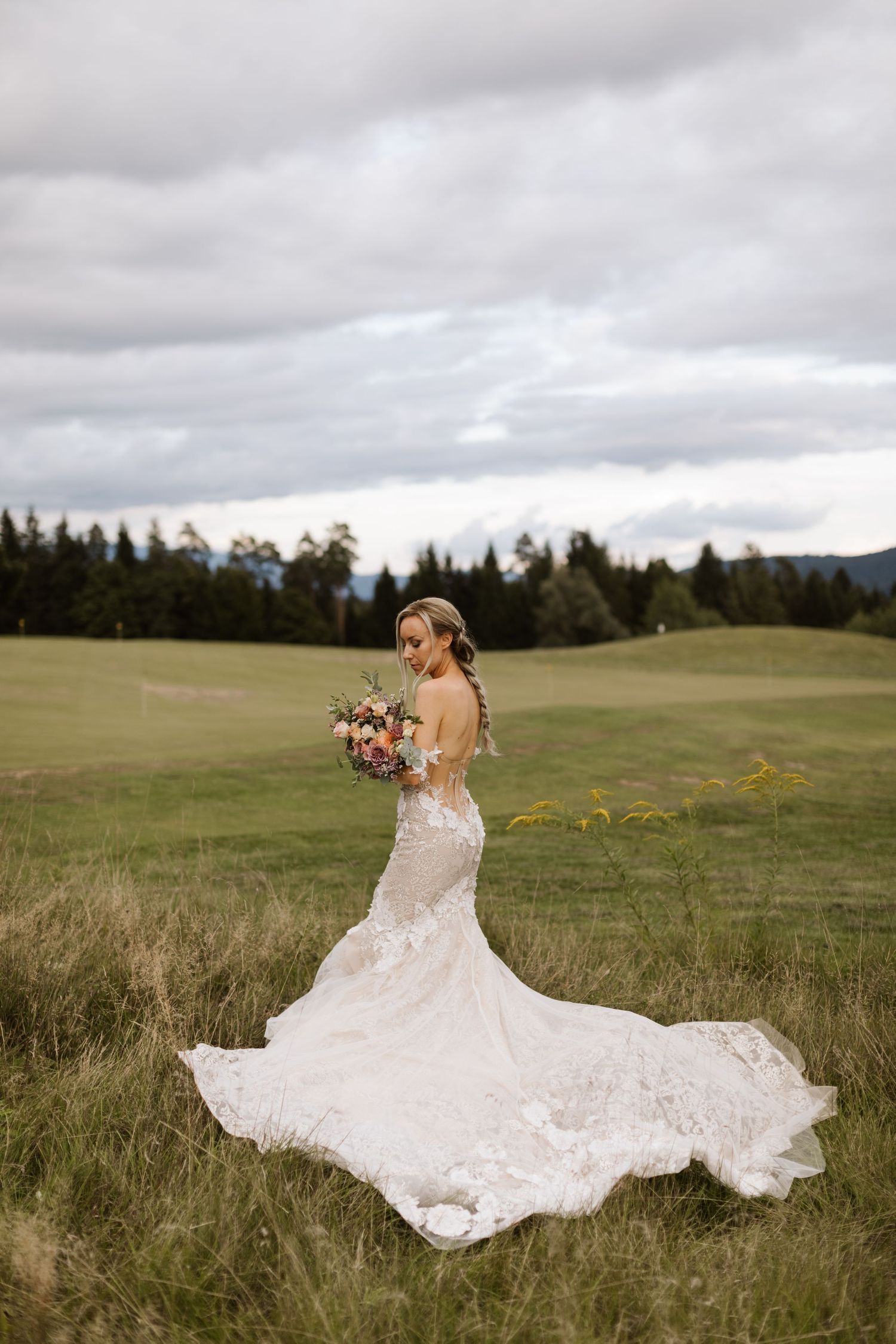 A bride in a flowing white lace dress twirls gracefully in a serene meadow with evergreen trees in the background.
